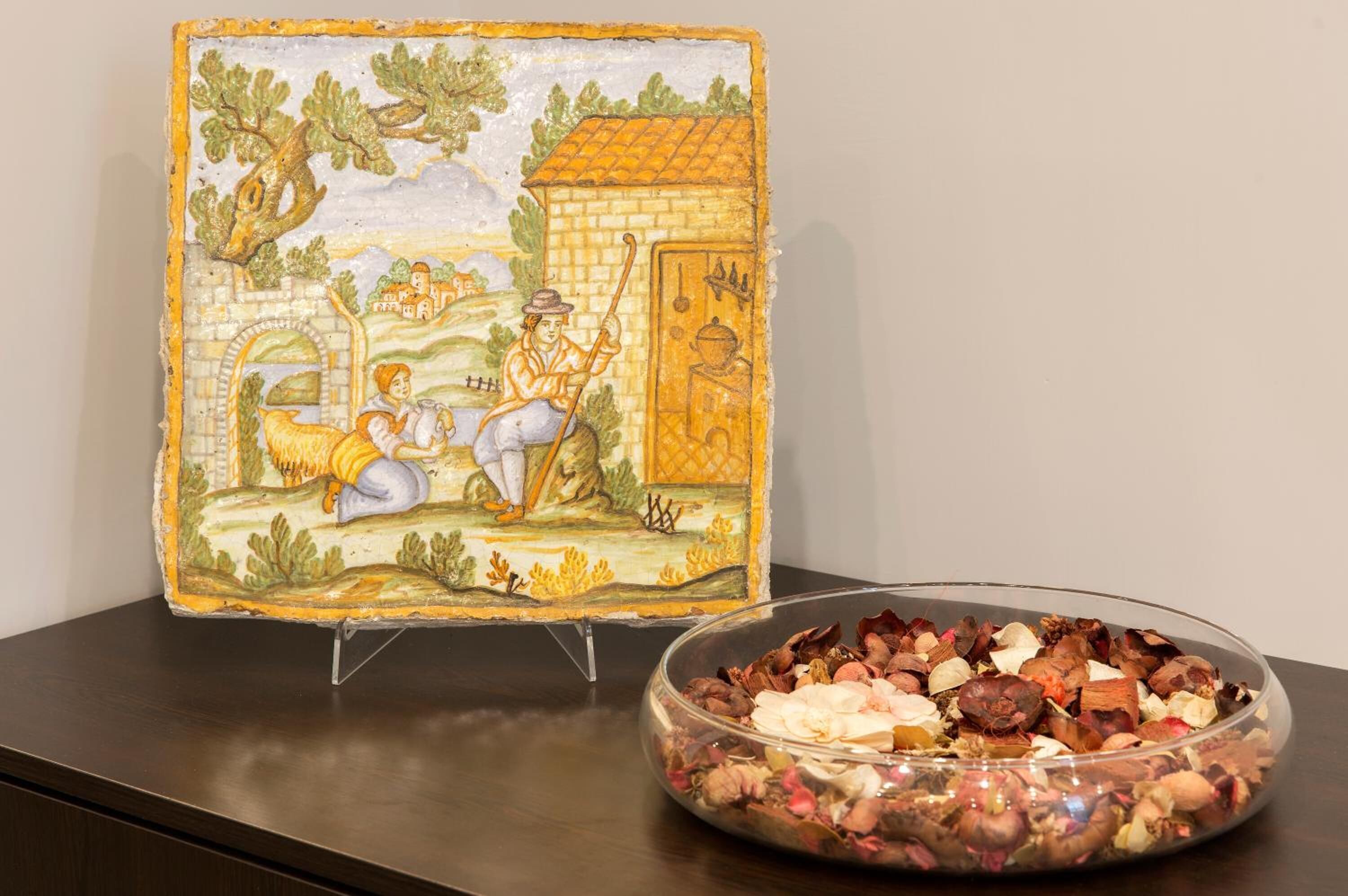A ceramic tile with a rural scene and a glass bowl filled with dried flowers.