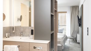 A modern hotel bathroom with a large mirror, a sink, and a vanity. There is a bed with a view of the outdoors.