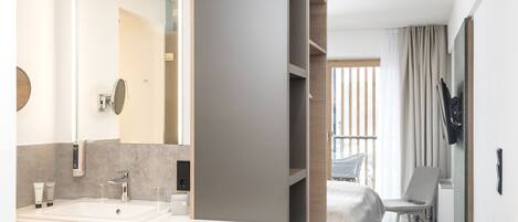 A modern hotel bathroom with a large mirror, a sink, and a vanity. There is a bed with a view of the outdoors.