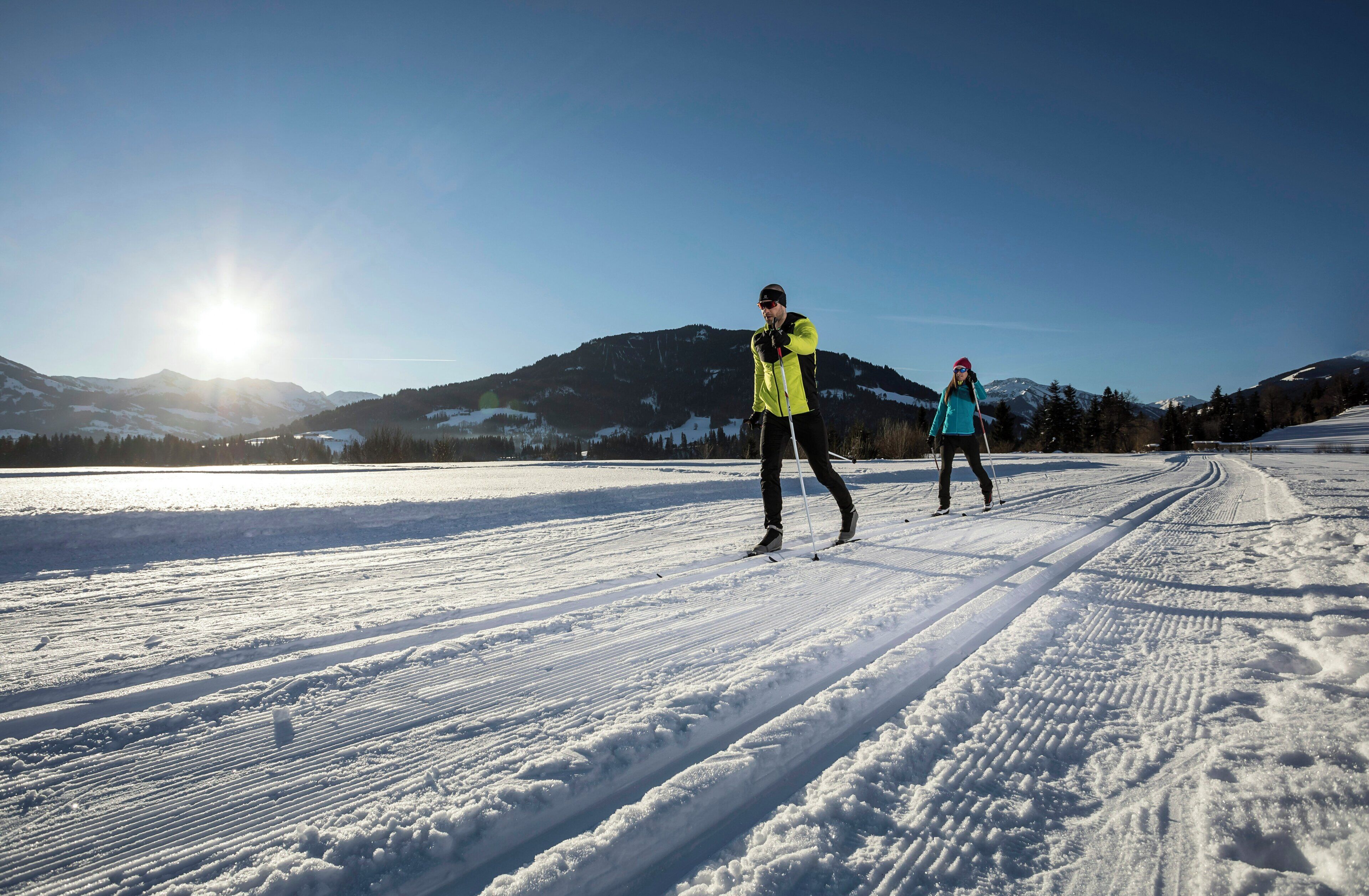 Two people cross-country skiing on a snow-covered path with mountains in the background.