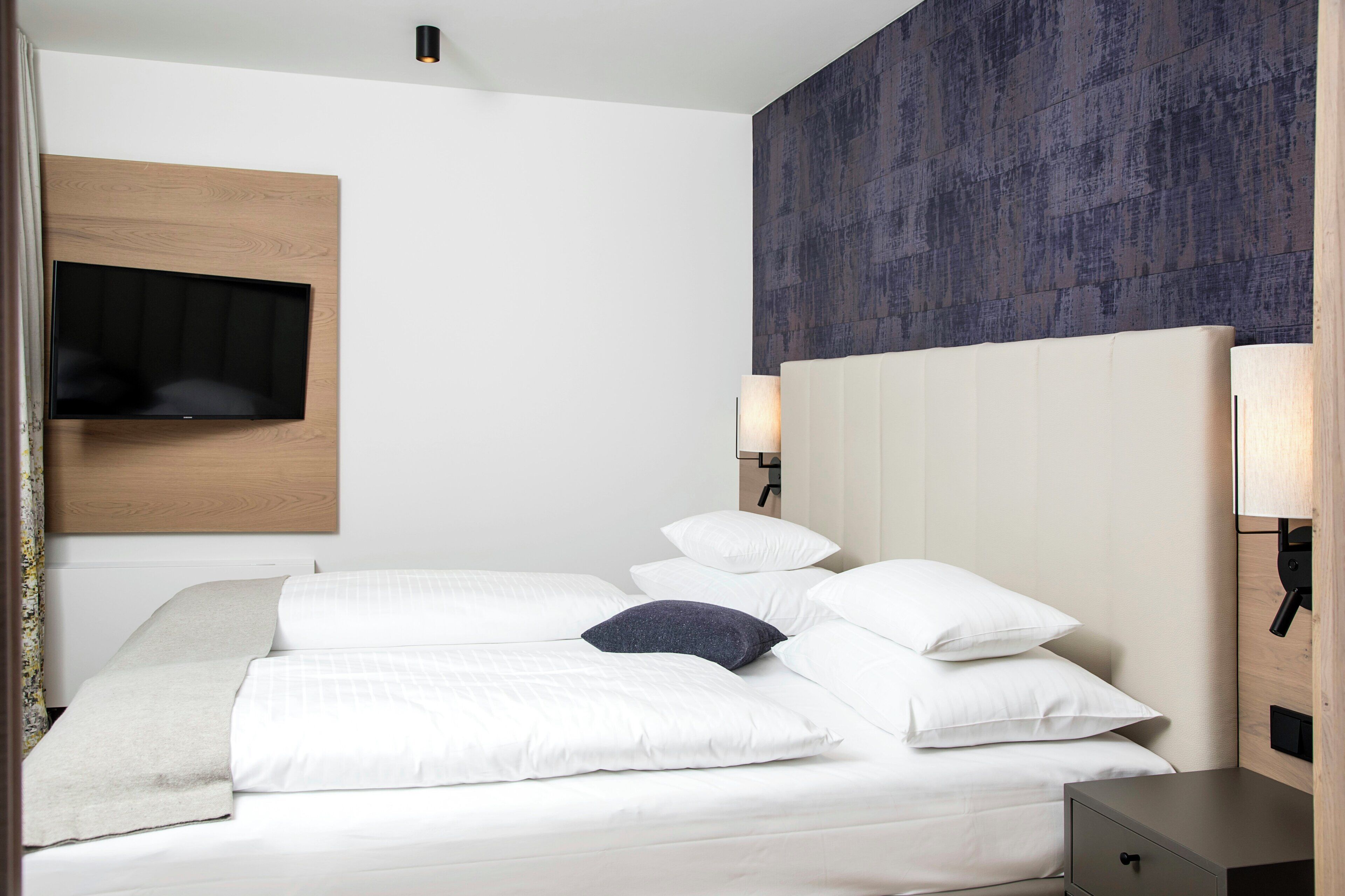 A hotel room with a bed, a flat-screen TV mounted on the wall, and a headboard with built-in lighting.
