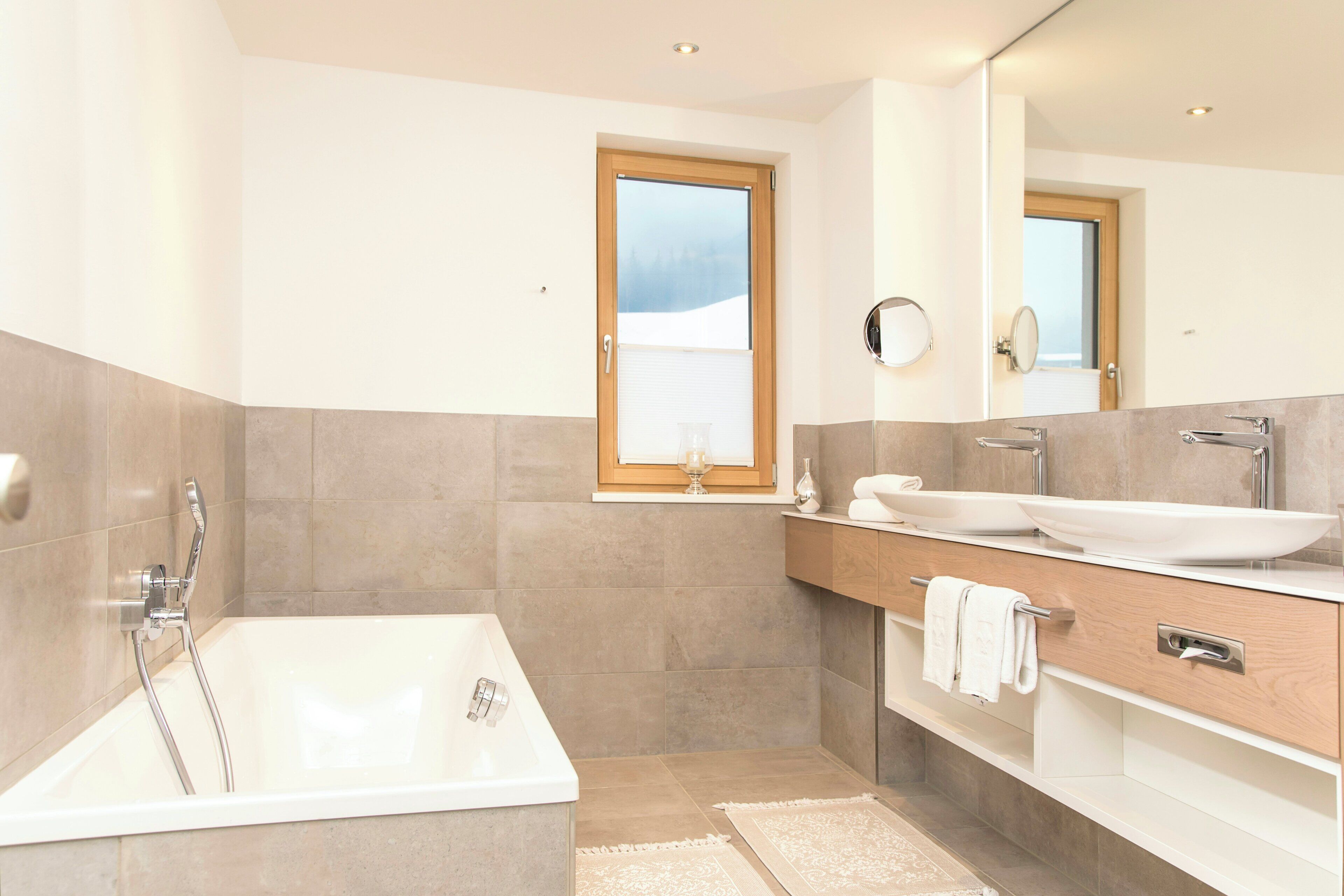 A modern bathroom with a bathtub, two sinks, a large mirror, and a window with a view.