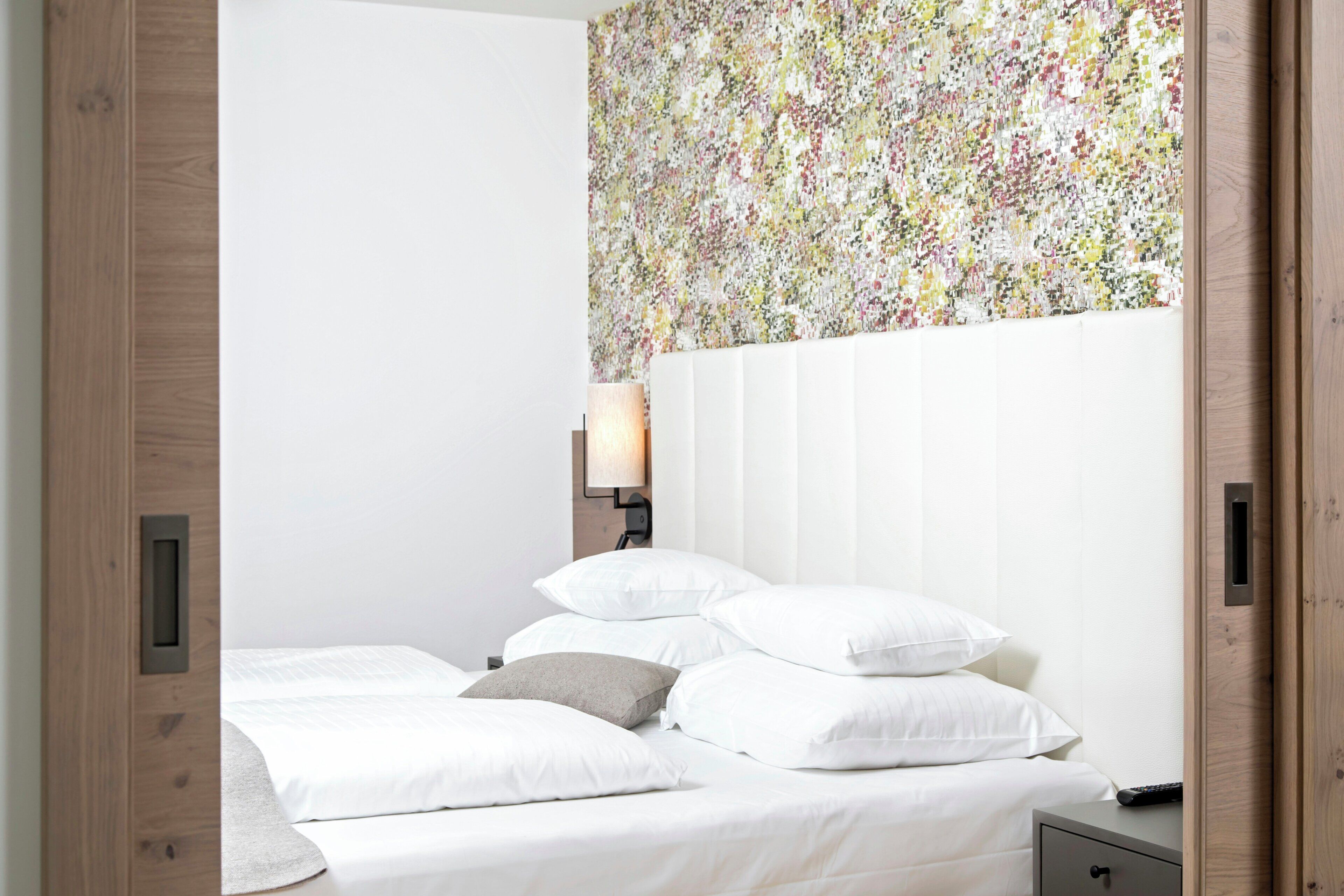 Un dormitorio con cama, almohadas, una mesita de noche, lámpara y papel tapiz con estampado floral.
