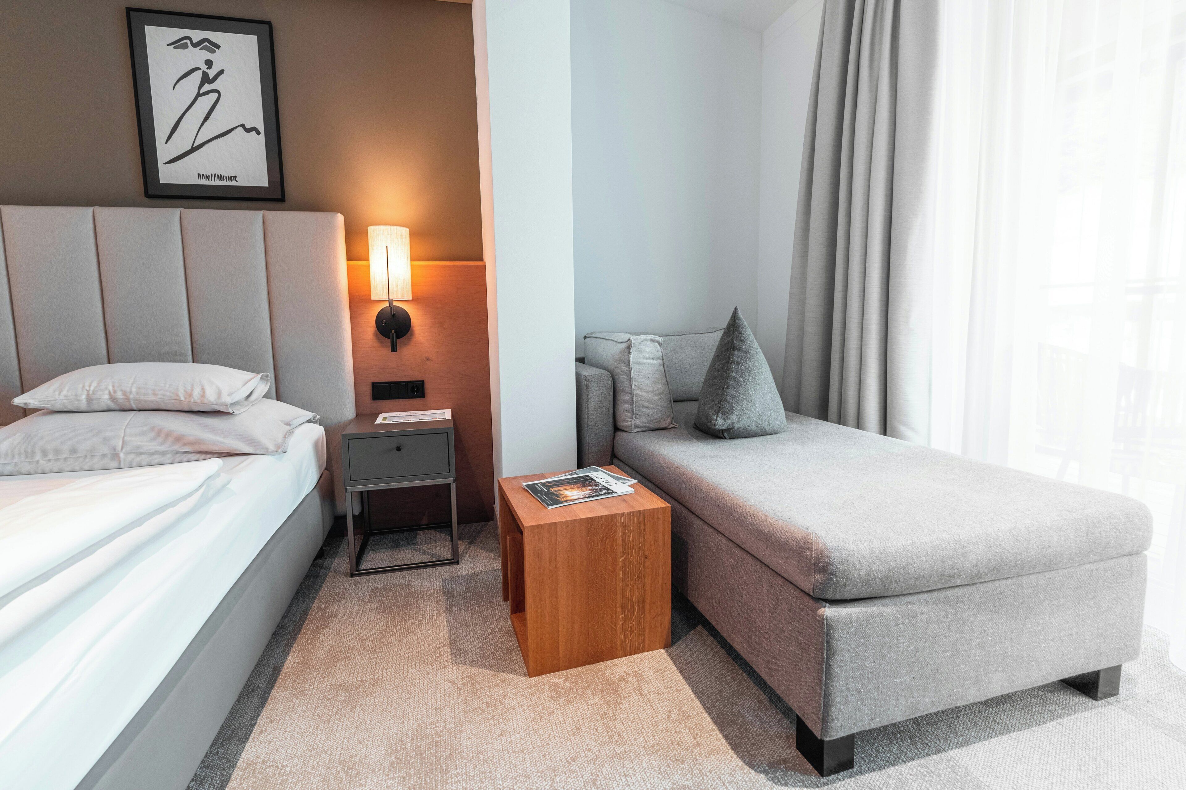 Habitació d'hotel amb llit, una petita taula de nit de fusta, taula de lectura, sofà i làmpada penjada a la paret.