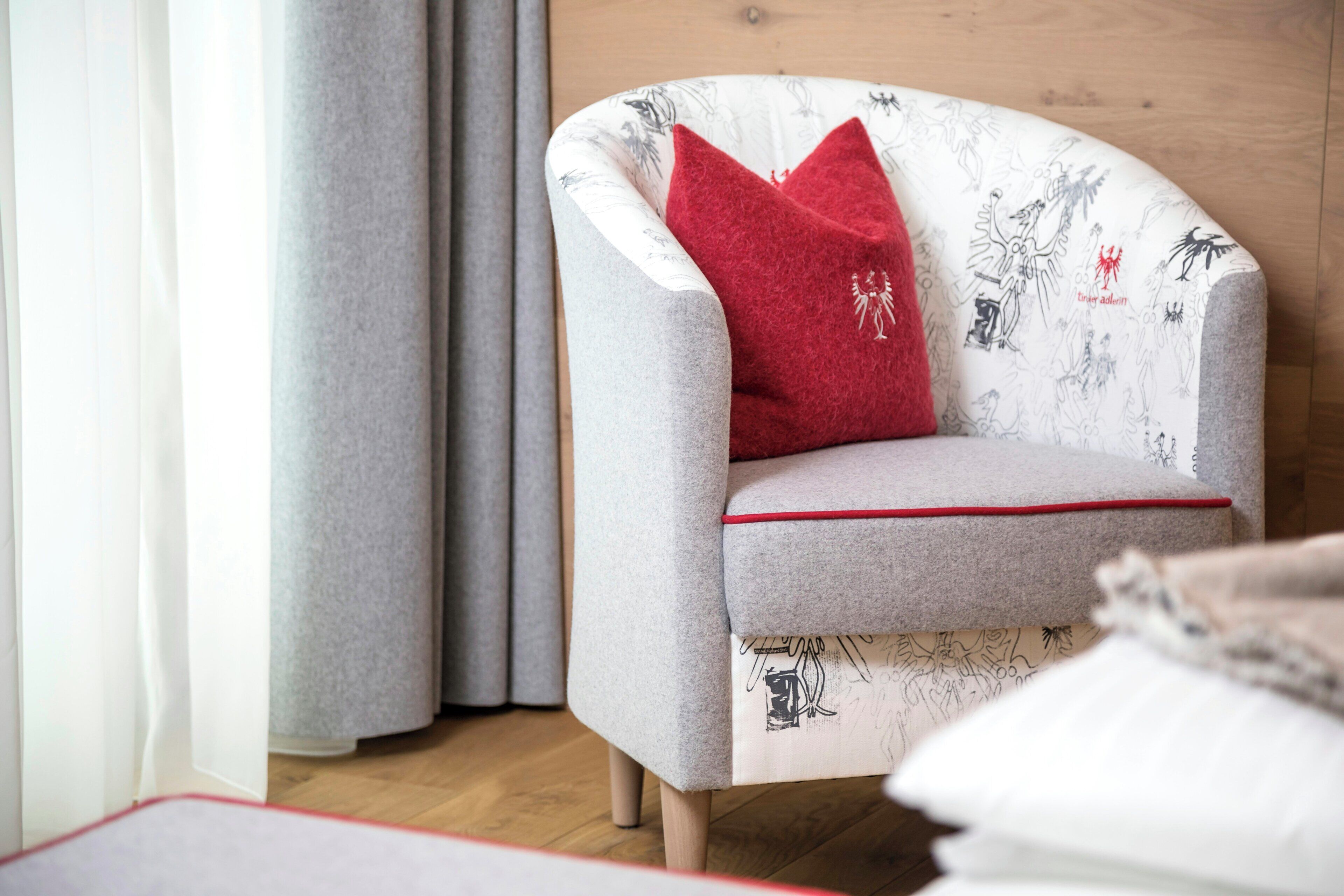 Un sillón moderno con un cojín rojo, ubicado en una habitación con piso de madera y una cortina blanca.