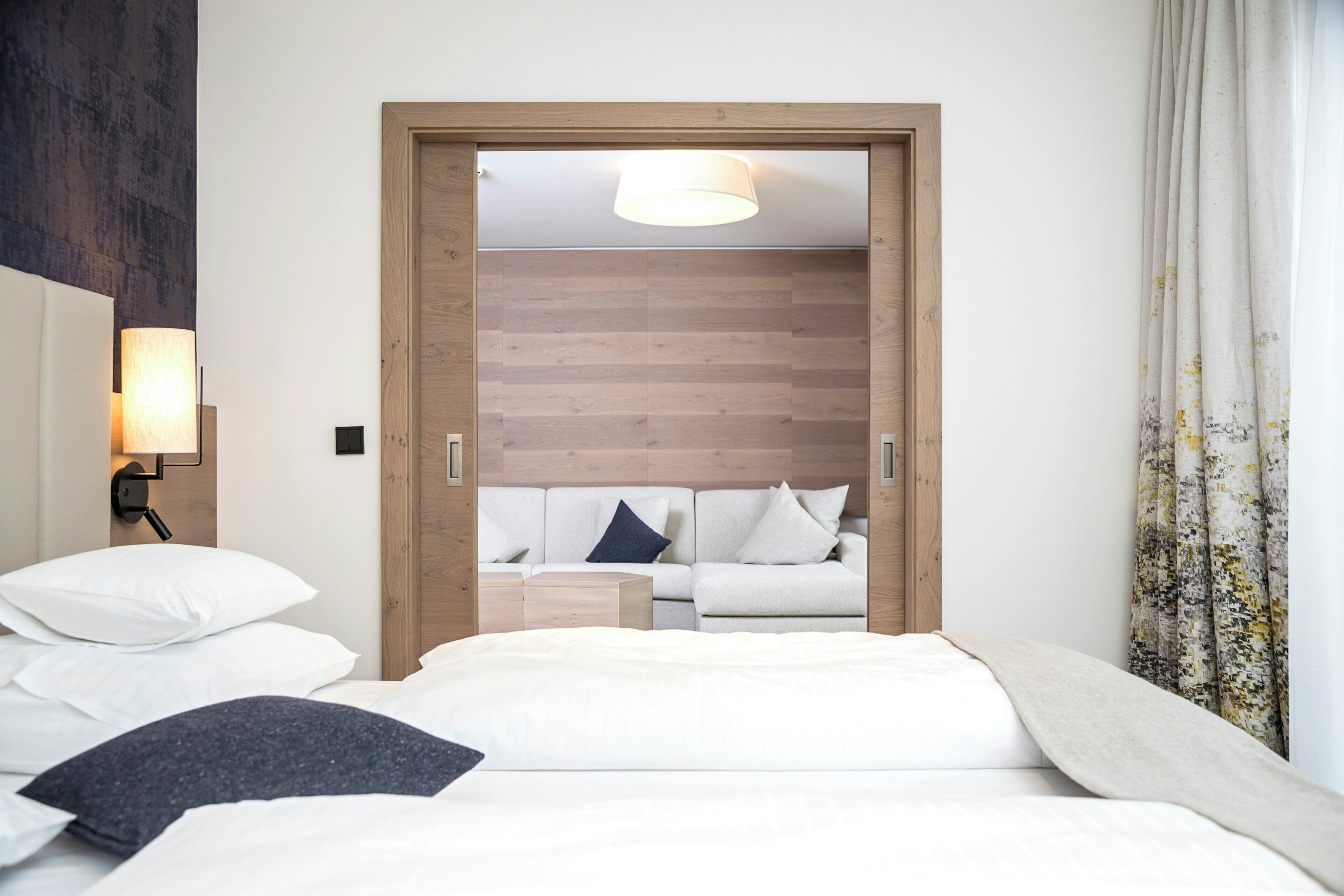 A hotel room with a bed, a nightstand, a lamp, and a view of a wooden wall and a sofa in the adjacent room.