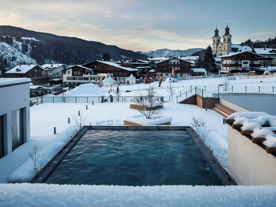 Zona exterior cubierta de nieve con piscina, bancos de madera y vista a un pueblo con edificios y montañas al fondo.