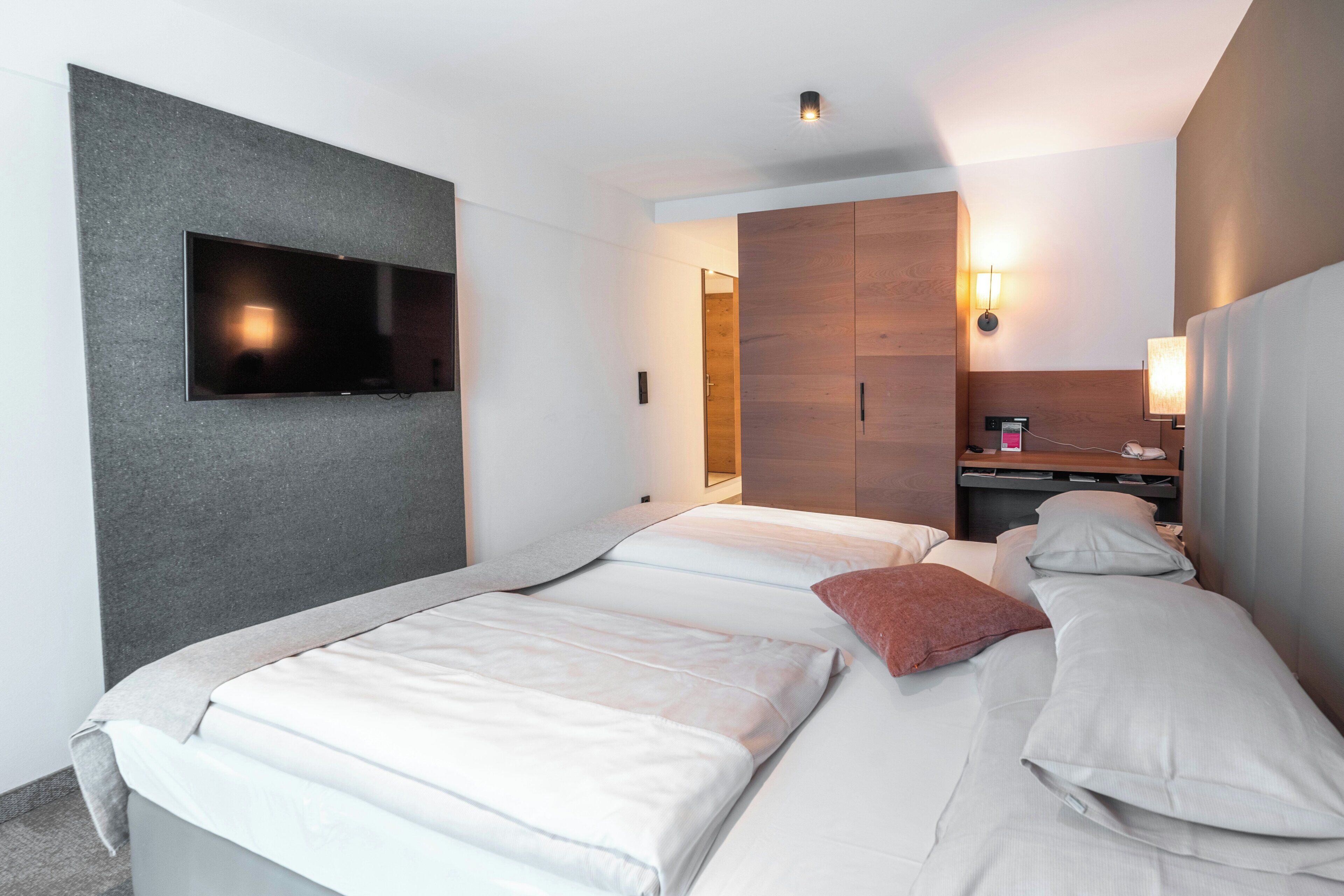 Une chambre d’hôtel avec deux lits, un téléviseur à écran plat fixé au mur, une armoire en bois et une table de chevet avec une lampe.