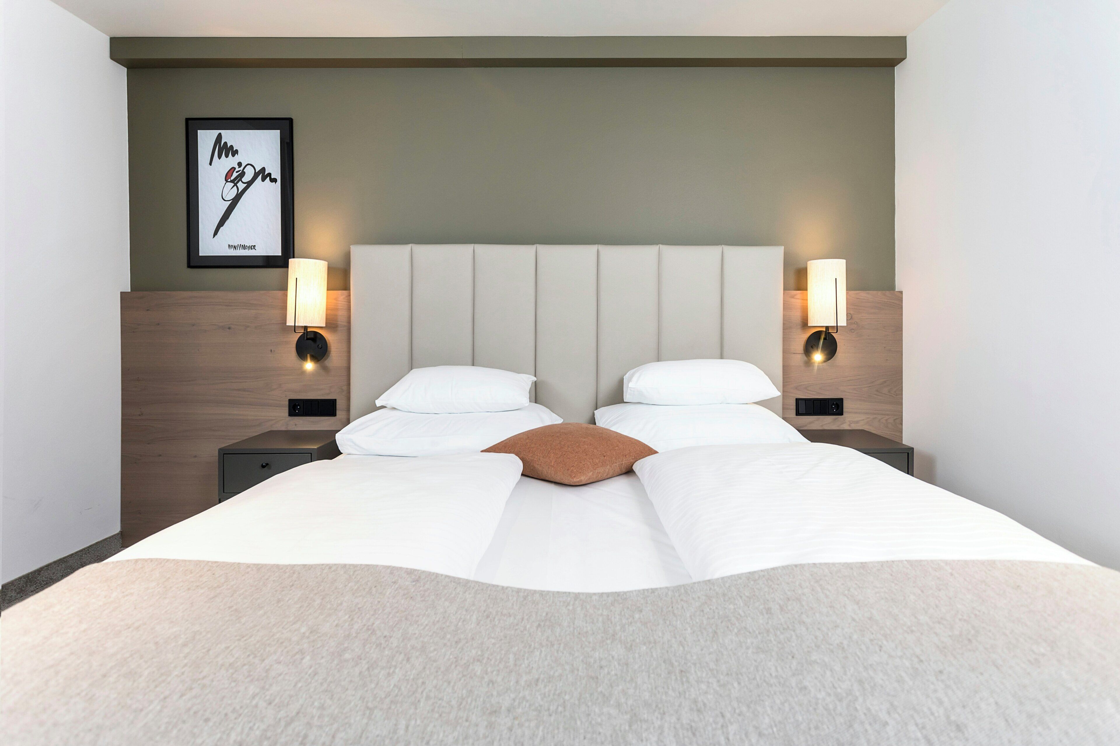 Habitación de hotel con una cama grande, dos mesitas de noche, lámparas fijadas a la pared y una obra de arte enmarcada sobre la cama.