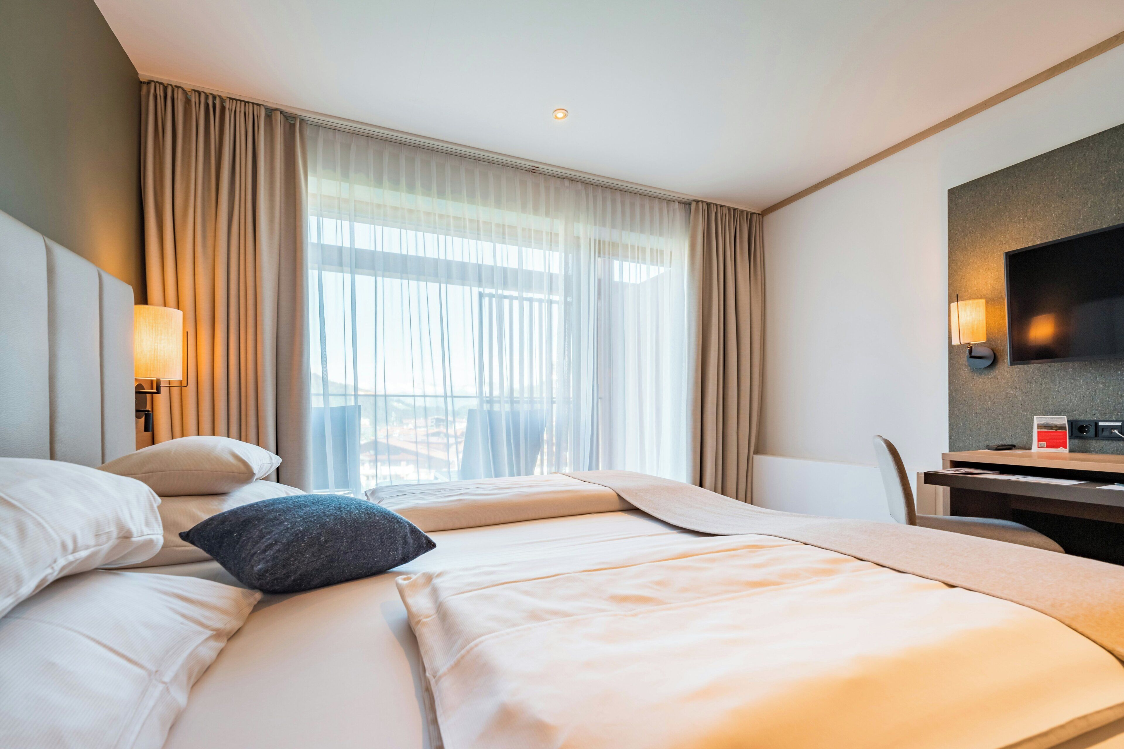Une chambre d’hôtel avec un lit, un bureau, une chaise, une télévision et une fenêtre avec des rideaux.