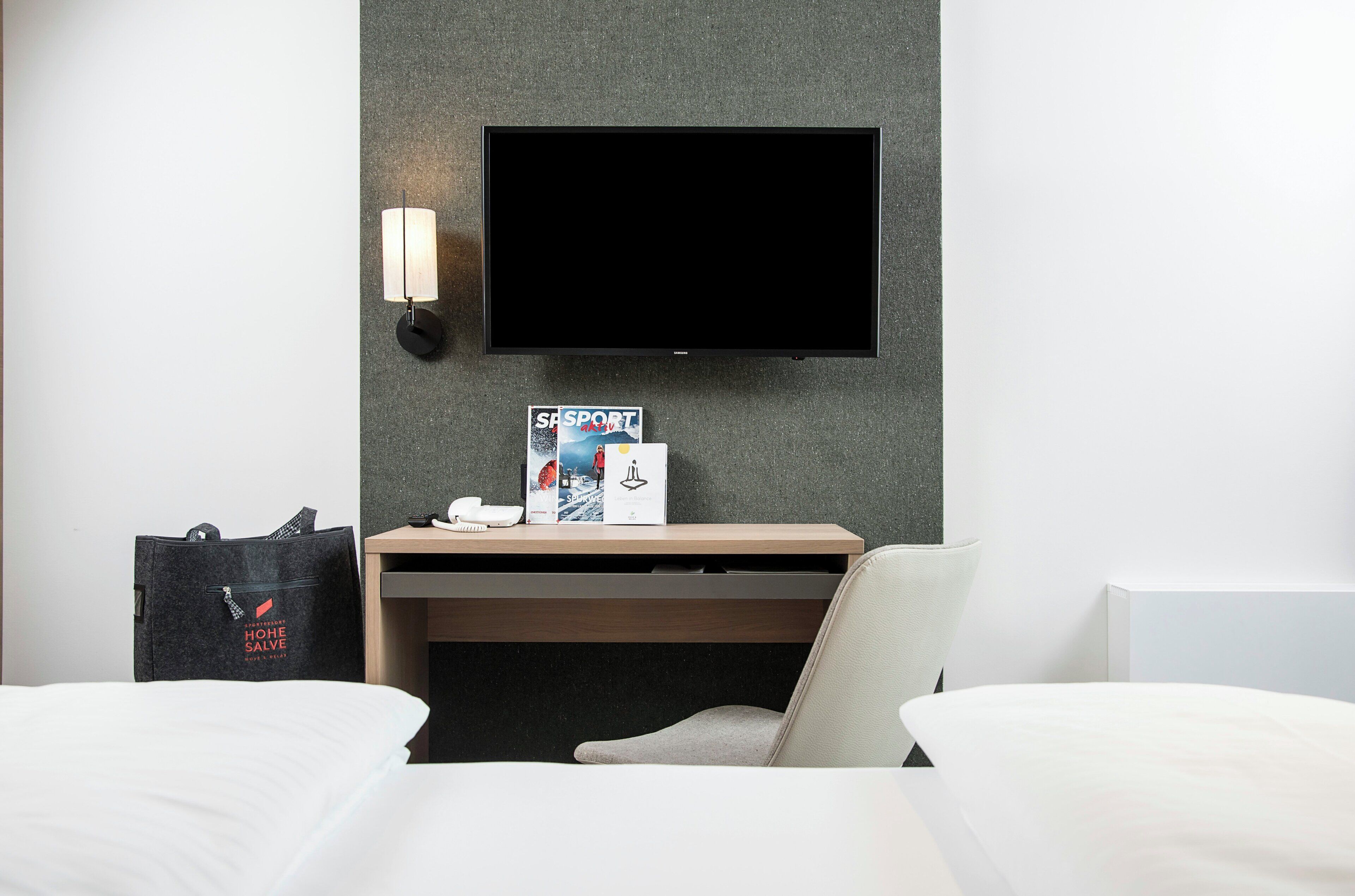 Ein Hotelzimmer mit einem Bett, einem Schreibtisch mit Flachbildfernseher, einer Wandlampe und einer Tasche auf dem Boden.