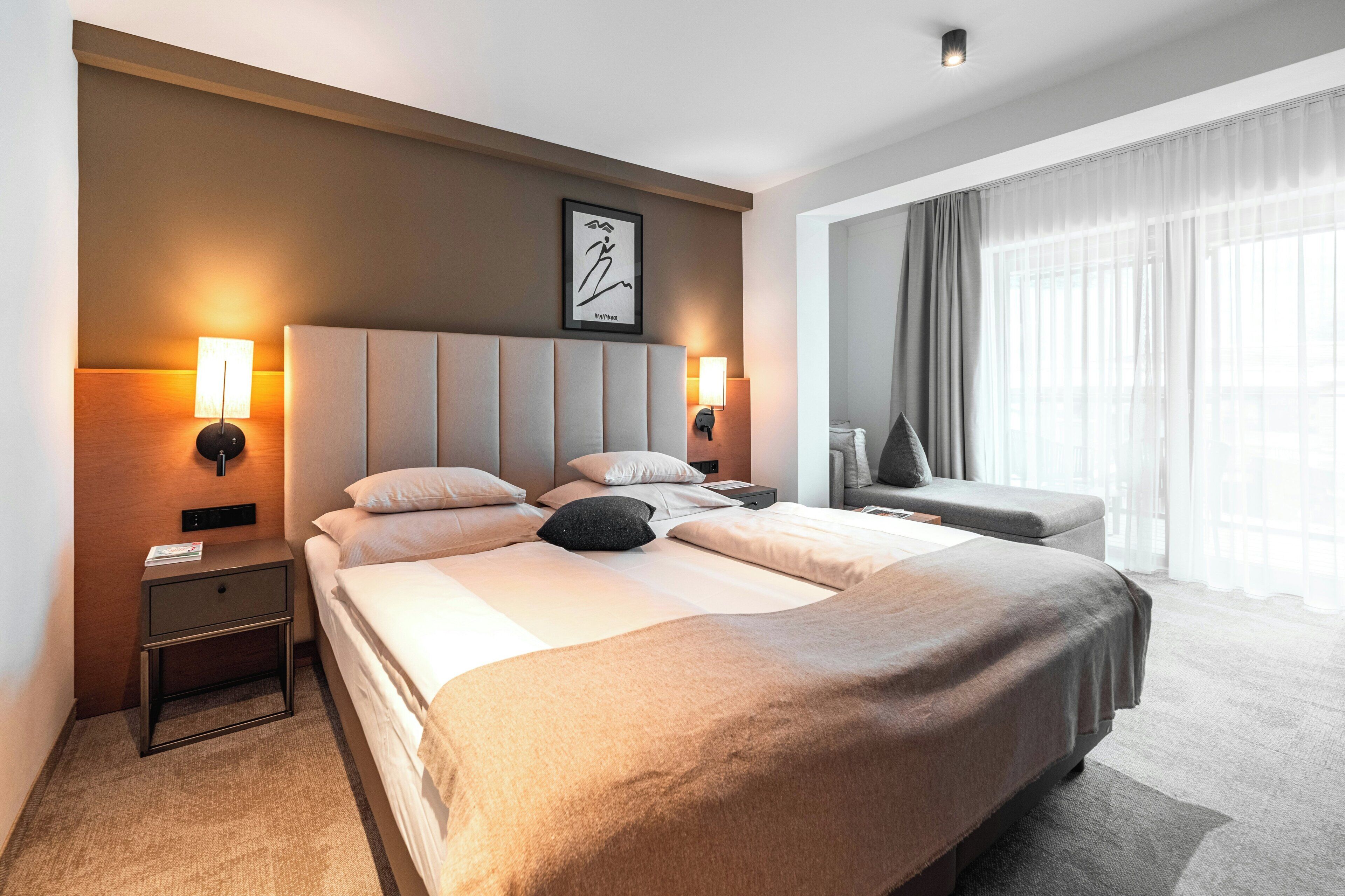 Ein modernes Hotelzimmer mit einem großen Bett, Nachttischlampen, einem Kopfteil, einem gerahmten Kunstwerk, einem Beistelltisch, einer Leselampe, einem Sofa und einem Fenster mit Vorhängen.