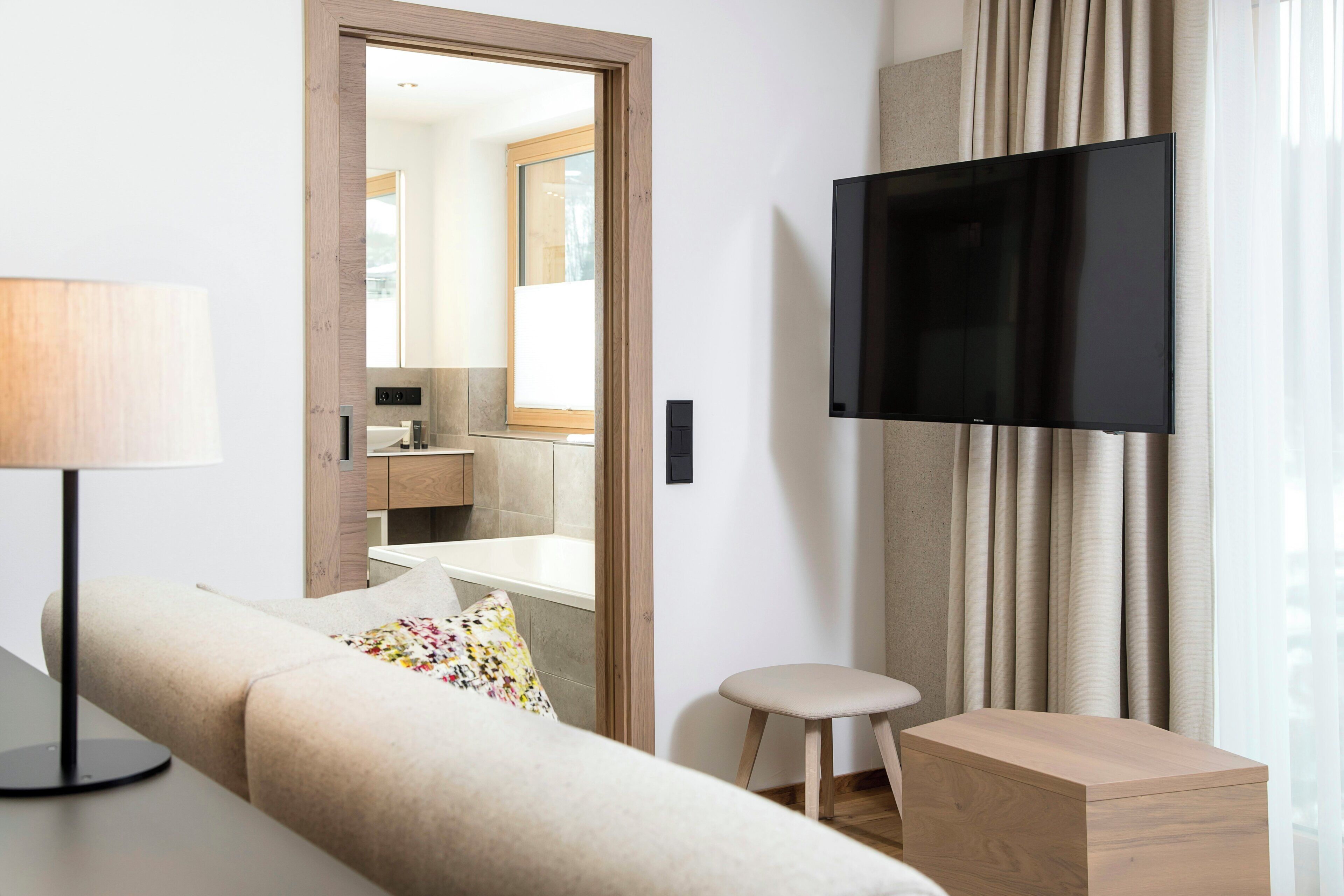 A hotel room with a sofa, a small stool, a TV, and a view of a bathroom through a doorway.