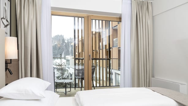 A hotel room with a bed, a nightstand, a lamp, and a view of a balcony with a table and chairs.