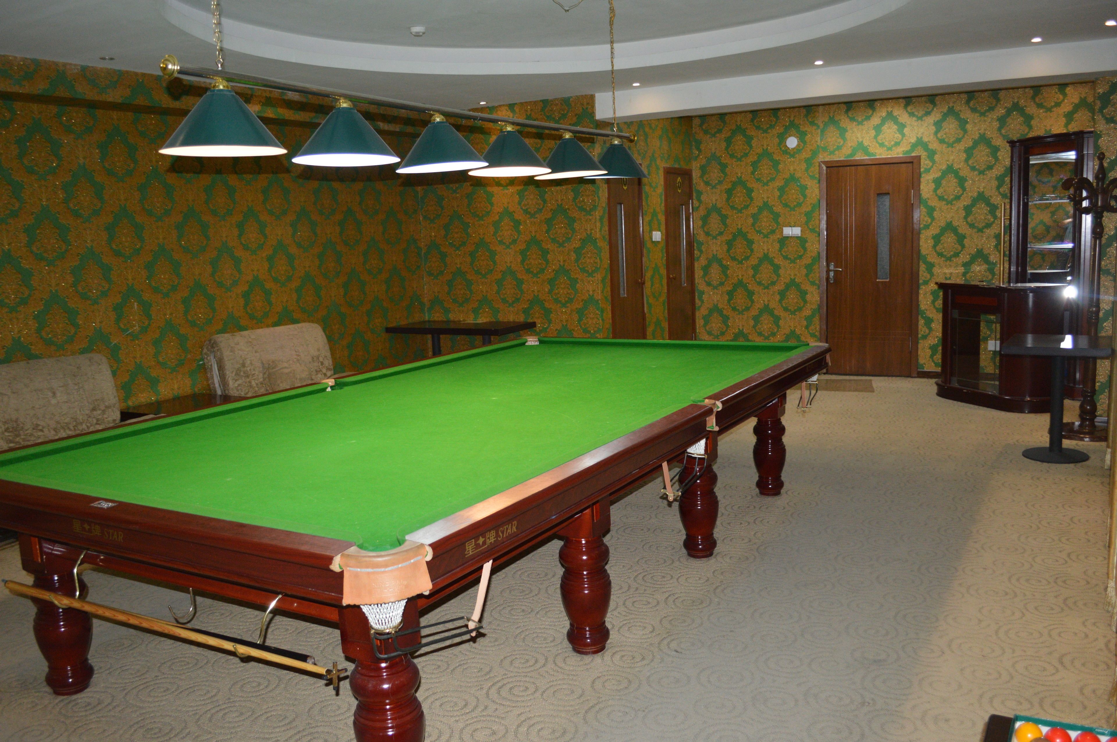 Een pooltafel met een groen vilt oppervlak, houten poten en een label met de tekst 'STAR'. Boven de tafel hangen vier groene pendelverlichting en er is een houten deur in de achtergrond zichtbaar.