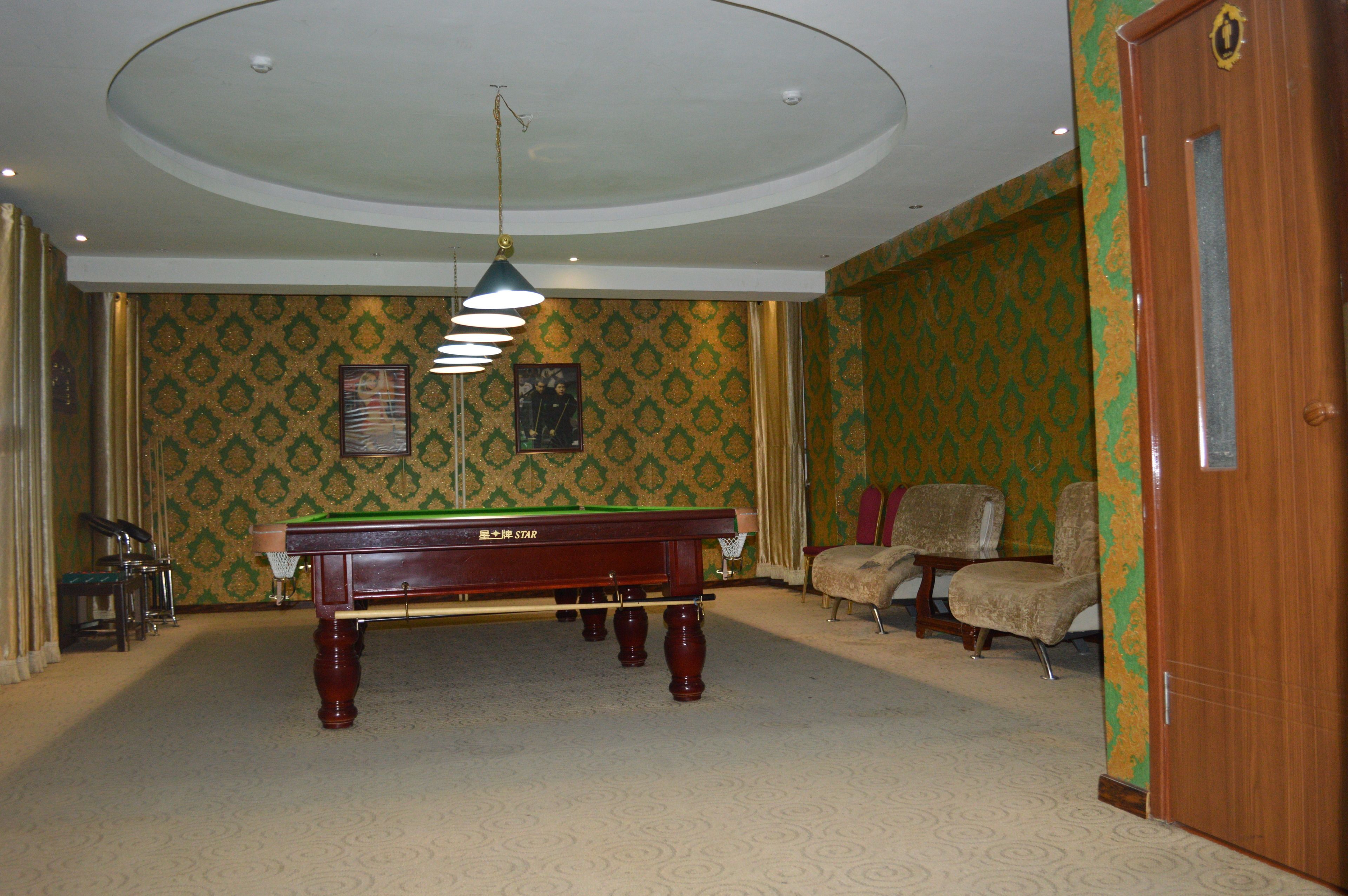 A room with a pool table, two armchairs, a ceiling light fixture, and patterned wallpaper.