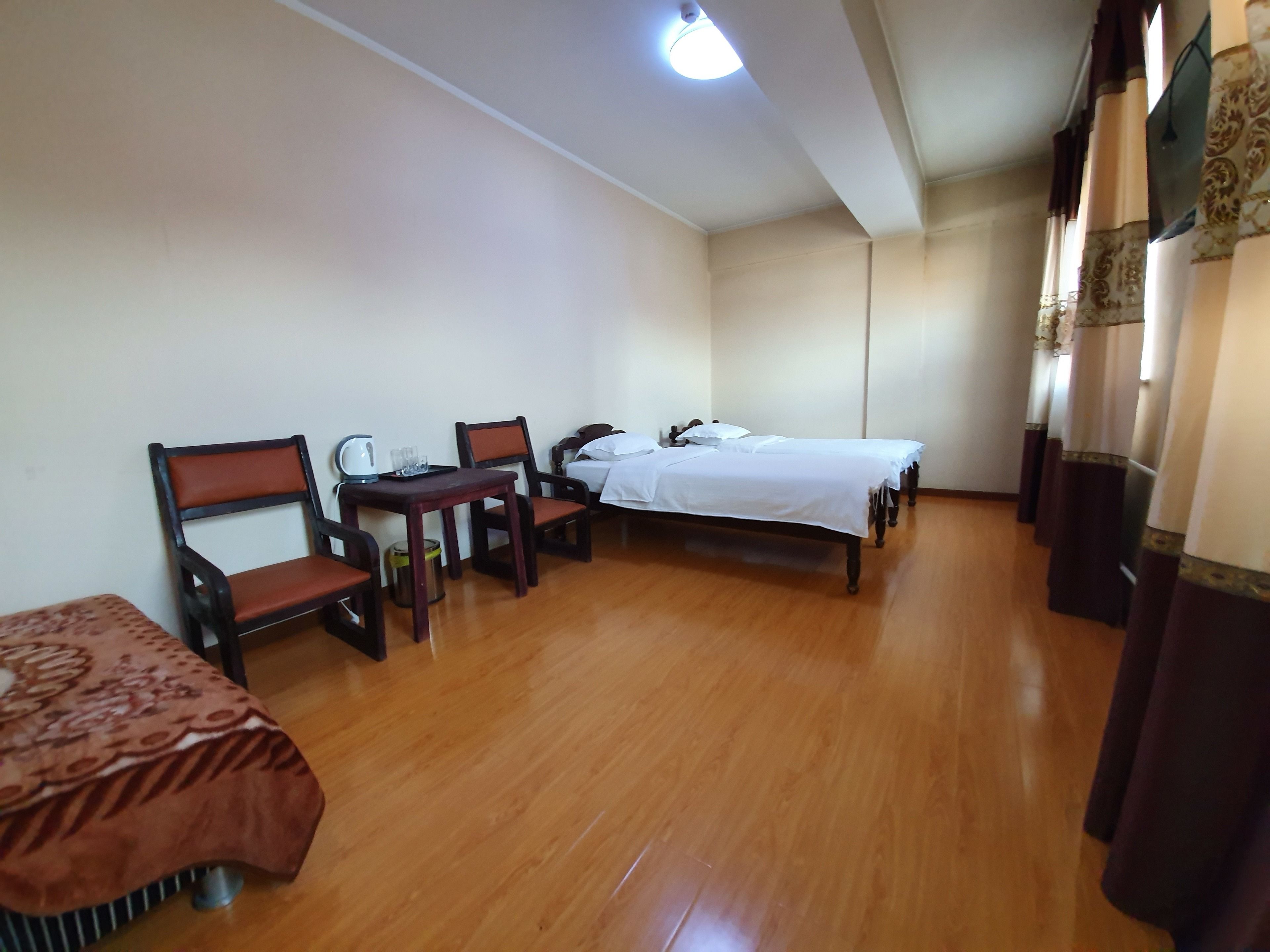 A hotel room with a wooden floor, a bed with white bedding, two wooden chairs, a small table with a kettle and glasses, and a patterned bedspread.