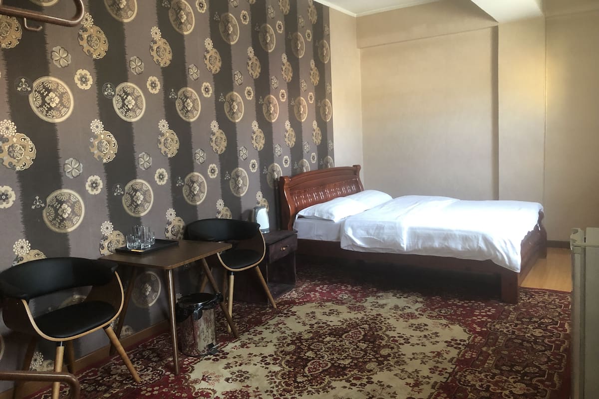 Quarto de hotel com uma cama, uma escrivaninha com duas cadeiras, um papel de parede estampado e um tapete vermelho.