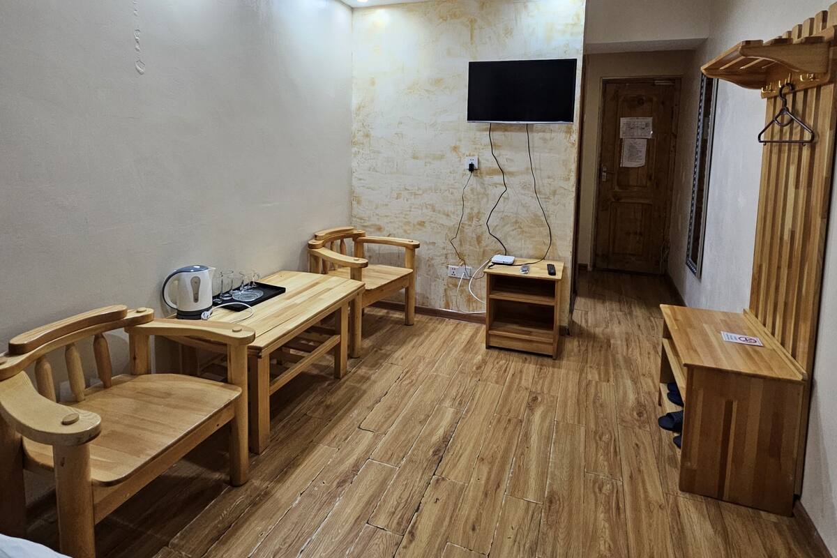 Quarto pequeno e acolhedor, com mesa e cadeiras de madeira, televisão na parede e porta-roupas de madeira.
