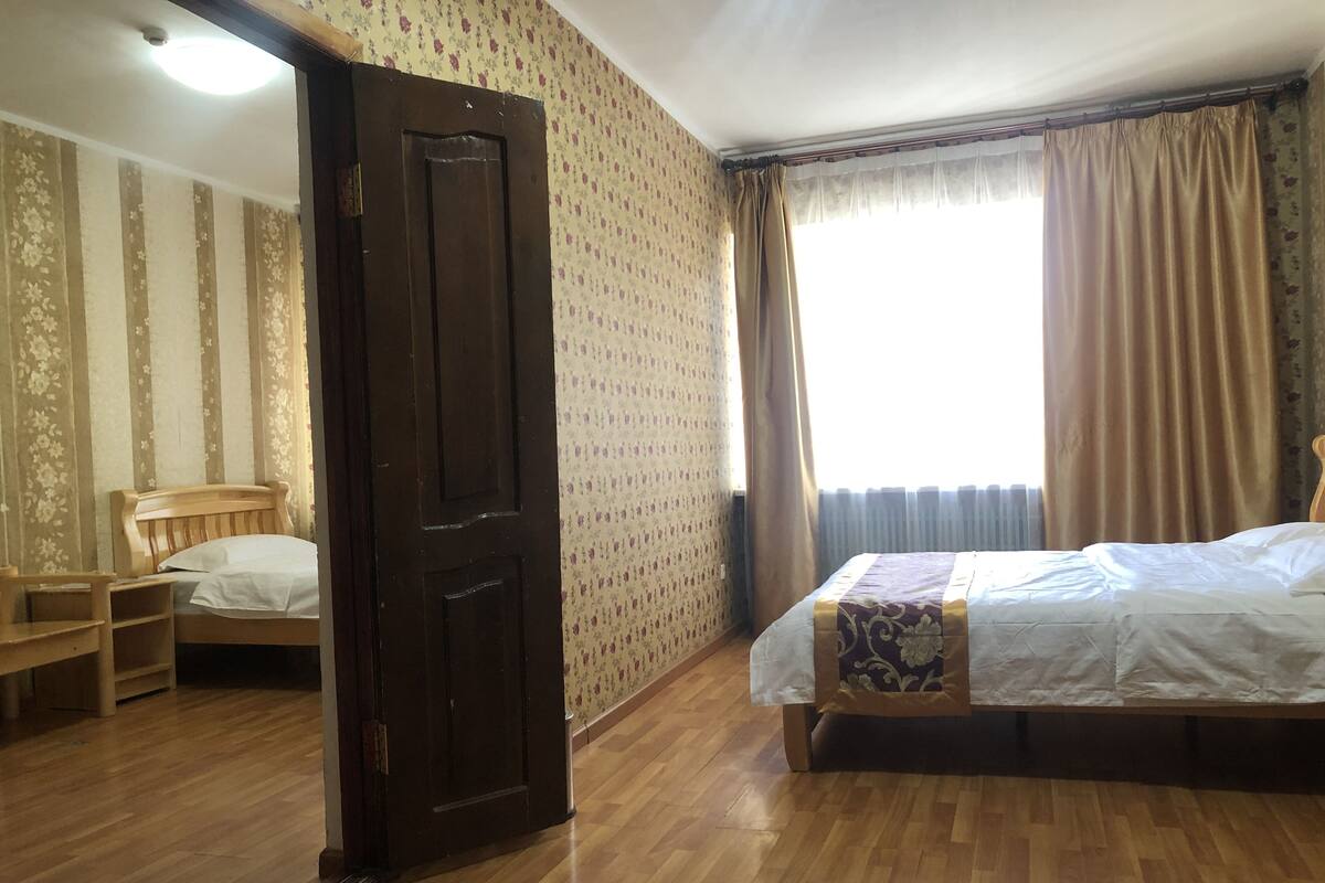 Um quarto com piso de madeira, uma cama com colcha estampada, uma cadeira e uma porta.