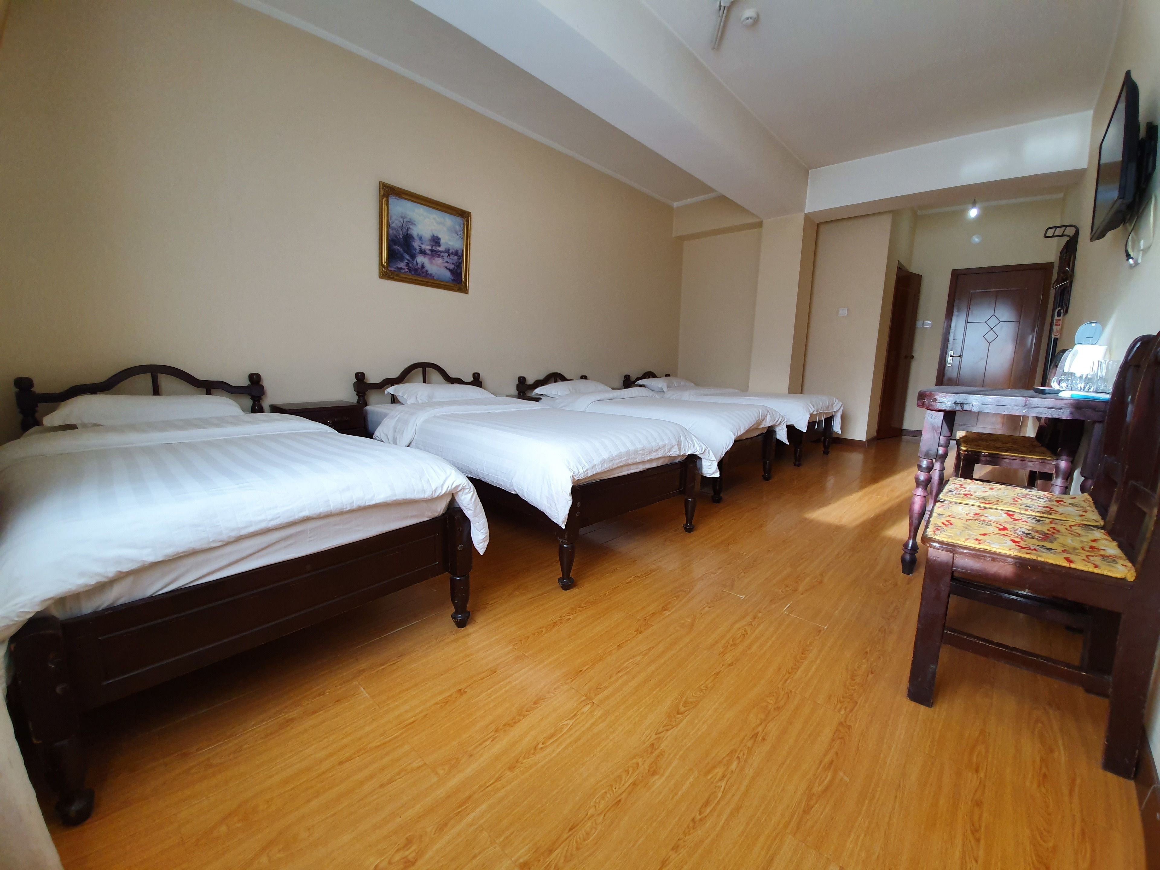 Habitación de hotel con cuatro camas de madera, una silla de madera, un escritorio con silla, un cuadro en la pared y una puerta de madera.