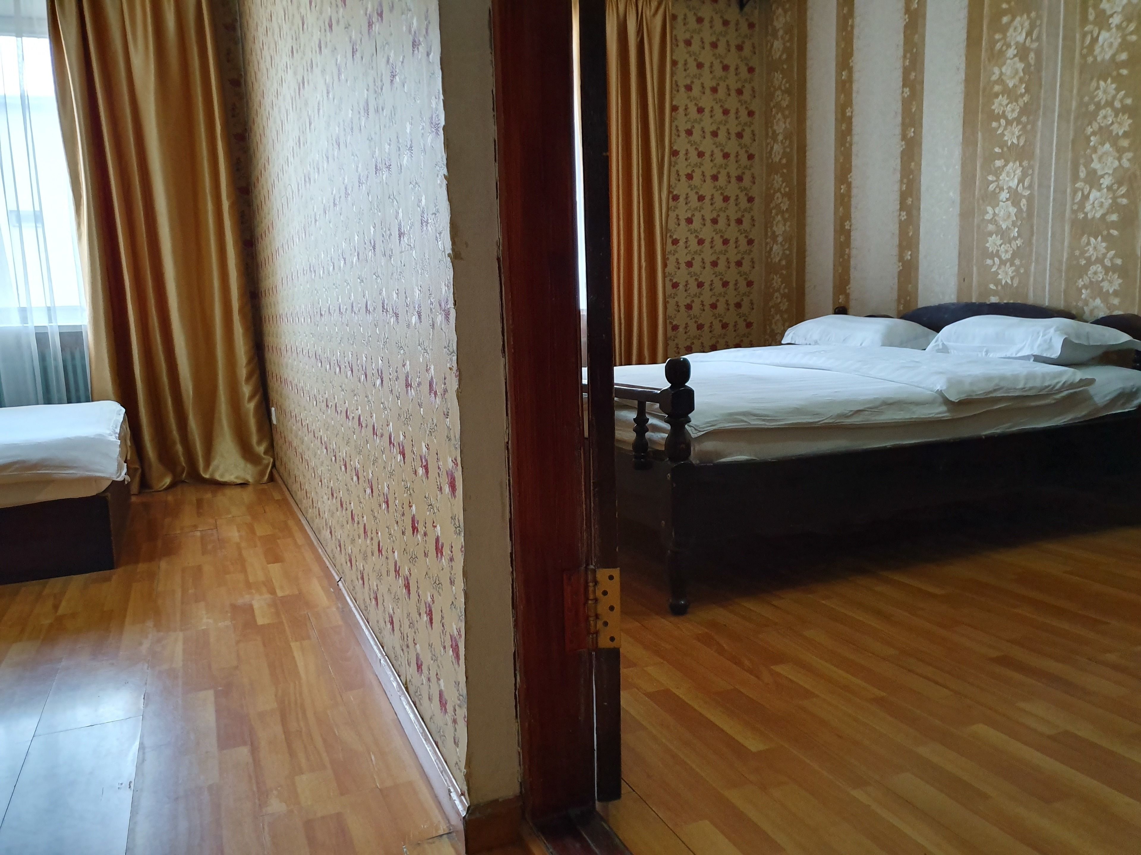 A bedroom with a wooden floor, a bed with white bedding, a nightstand, and a window with curtains.