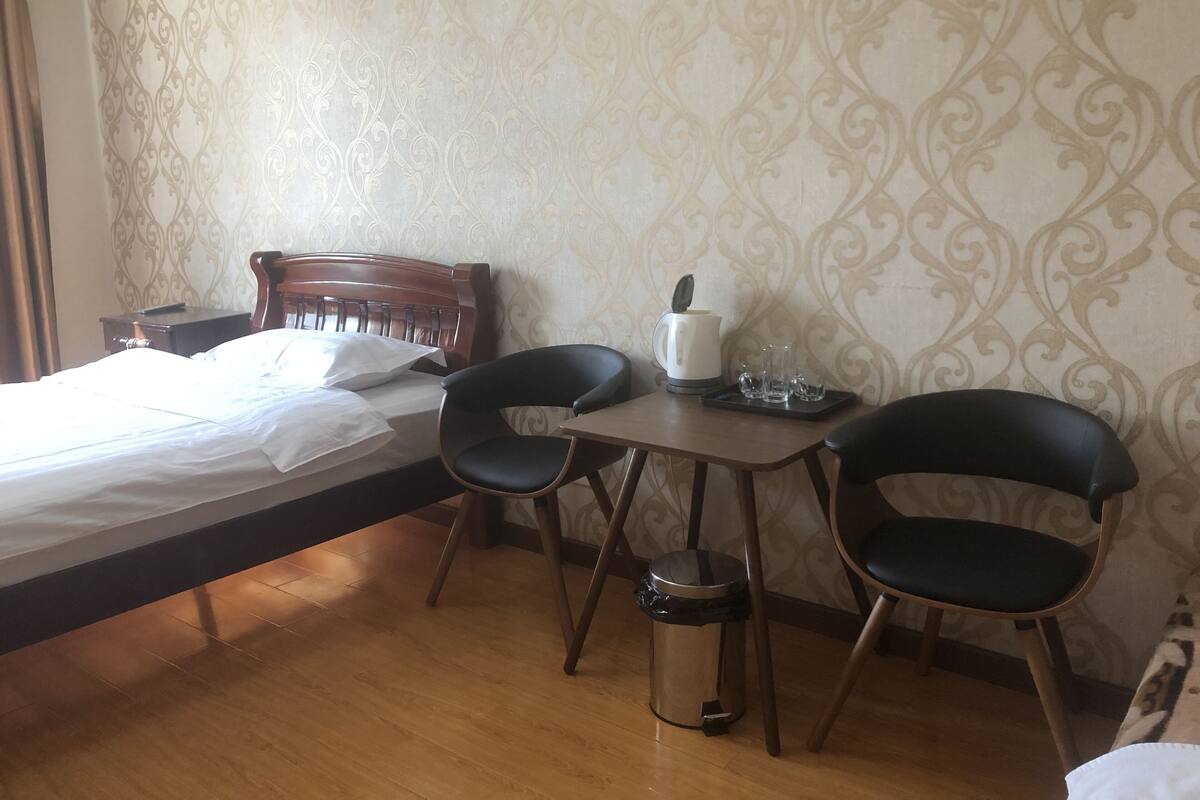Quarto de hotel com cama, duas cadeiras, uma mesinha com chaleira e copos, e um cesto de lixo.