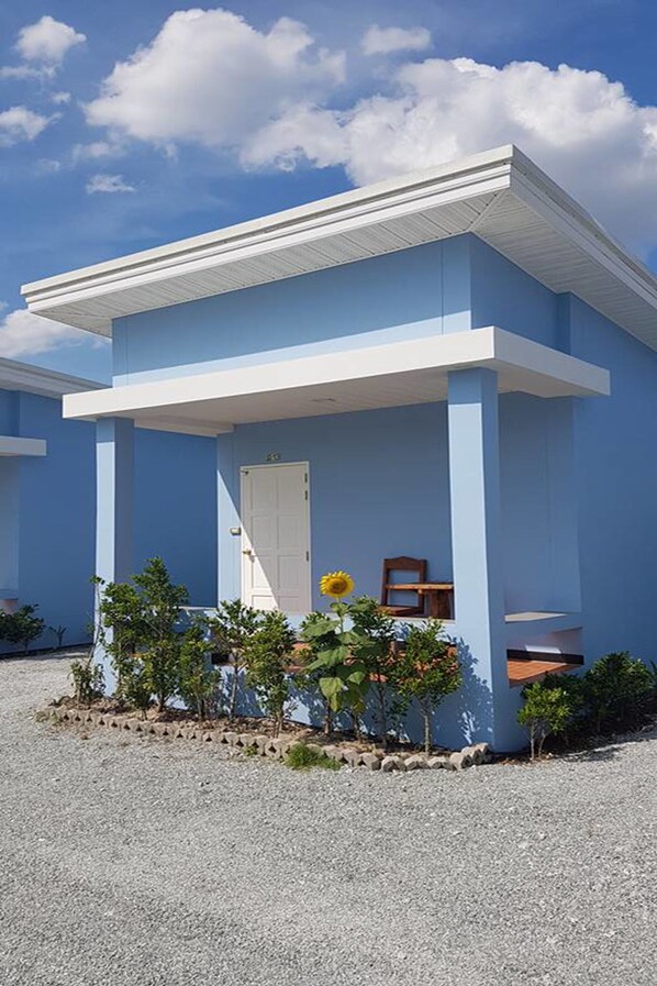 A single-story building with a flat roof and a covered porch. There is a door on the front and a small garden area with plants and a sunflower.