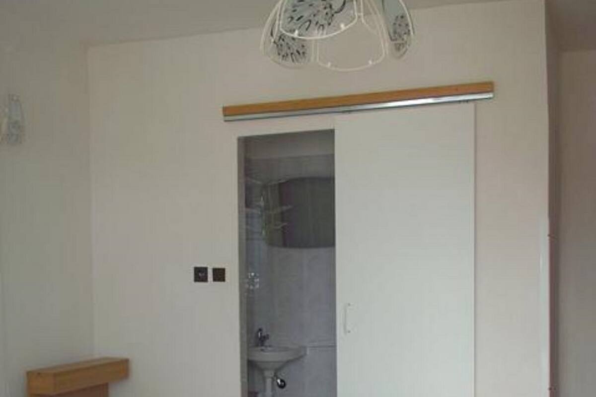 A bedroom with two beds, a wardrobe, a small bedside table, a hanging light fixture, and a red patterned rug.