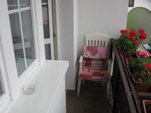 A small balcony with a white chair, a potted plant with red flowers, and a window with blinds. - Penzión Závada (Nova Lesna)