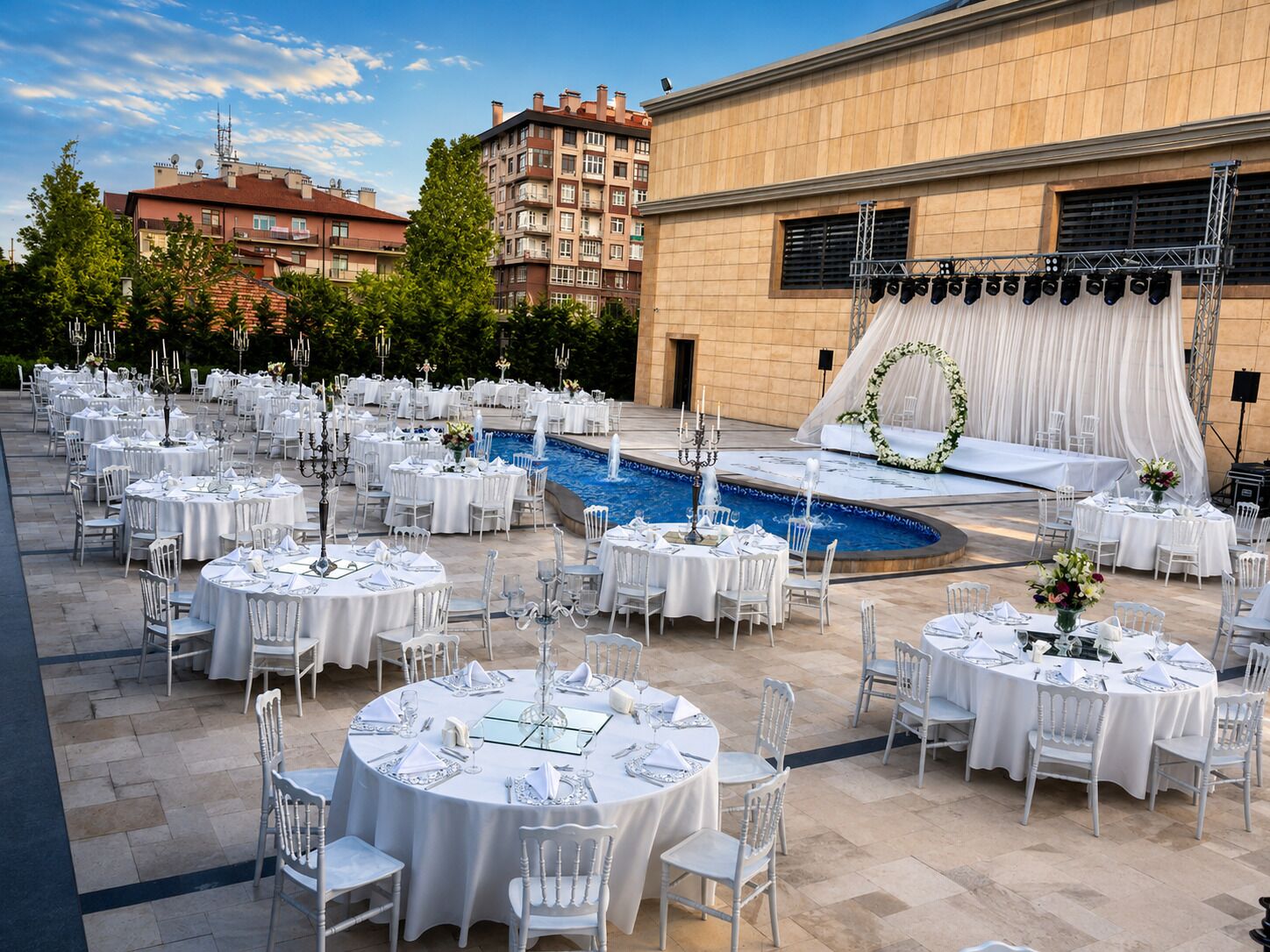 An outdoor wedding venue with round tables, chairs, a pool, and a stage with drapes.