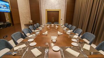 Un salón de conferencias con una larga mesa dispuesta para una reunión, equipado con sillas, platos, cubiertos y una pantalla de televisión en la pared.