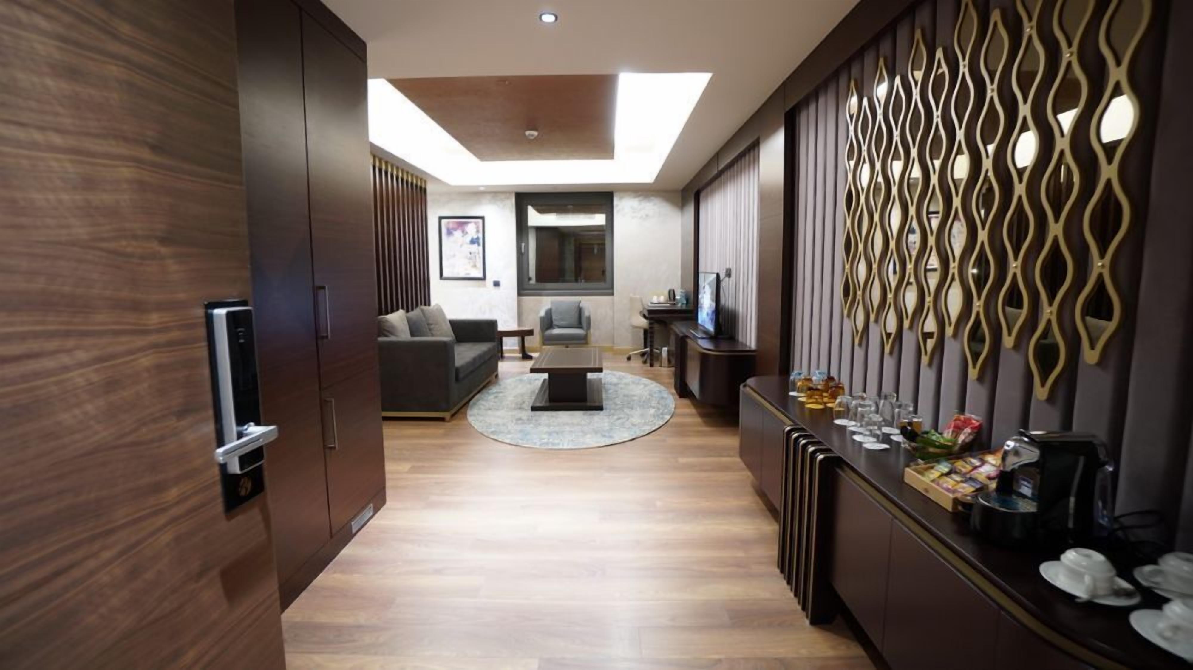 A modern living room with a sofa, coffee table, and a TV. There is a decorative wall with a geometric pattern and a small bar area with glasses and a coffee machine.