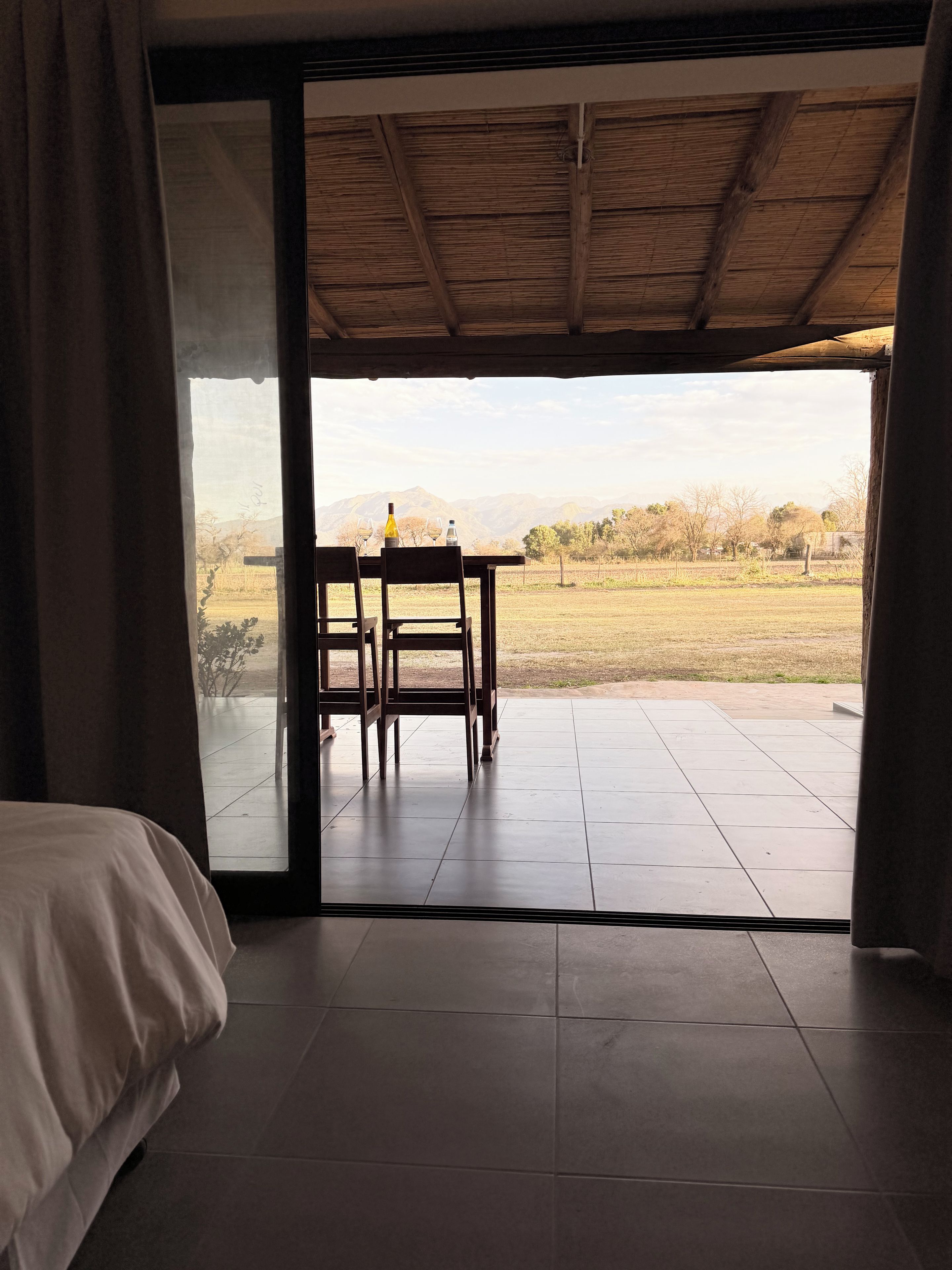 A room with a bed, a table with chairs, and a view of a field and mountains.