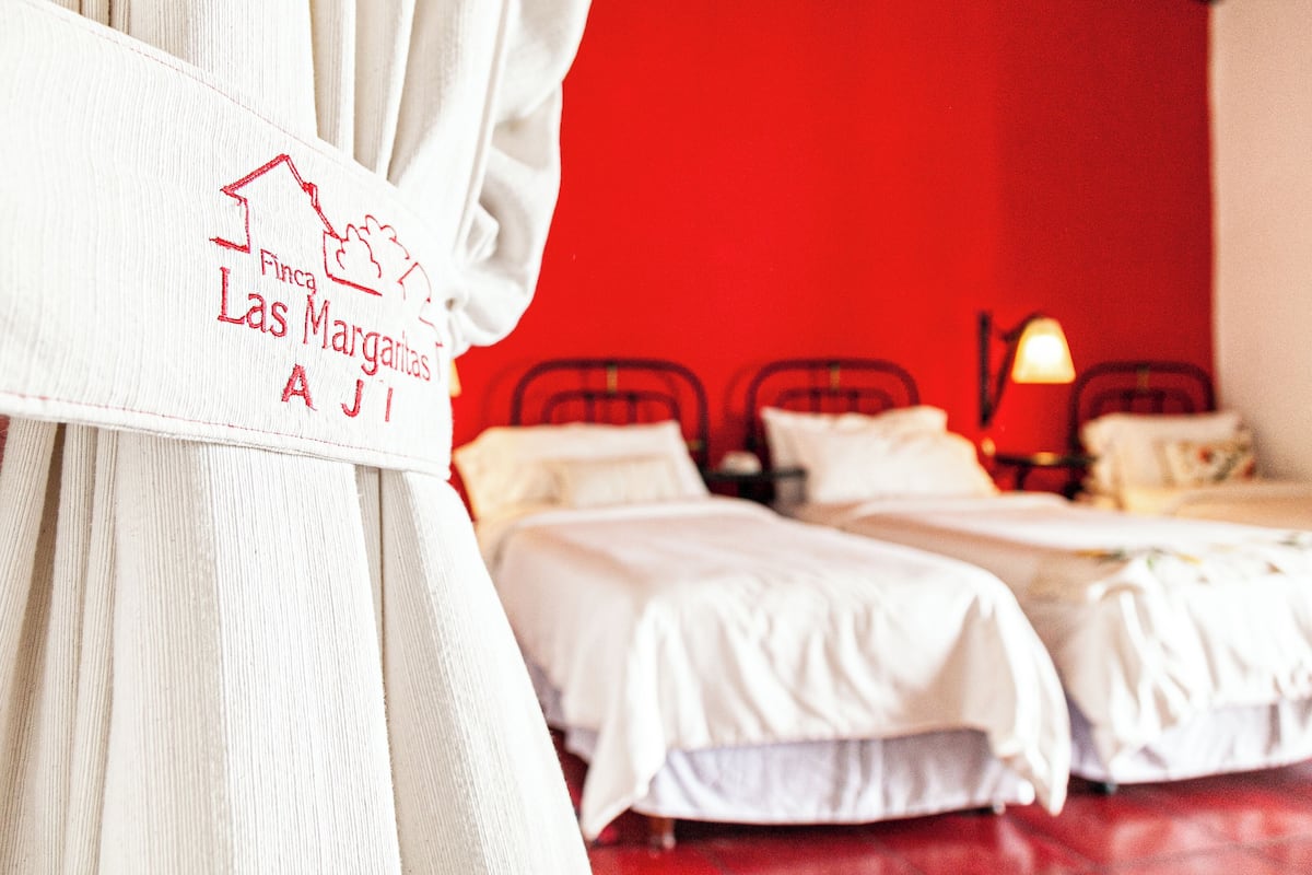 Ett hotellrum med två sängar, en röd vägg och en skylt på gardinen med texten "Finca Las Margaritas AJI".
