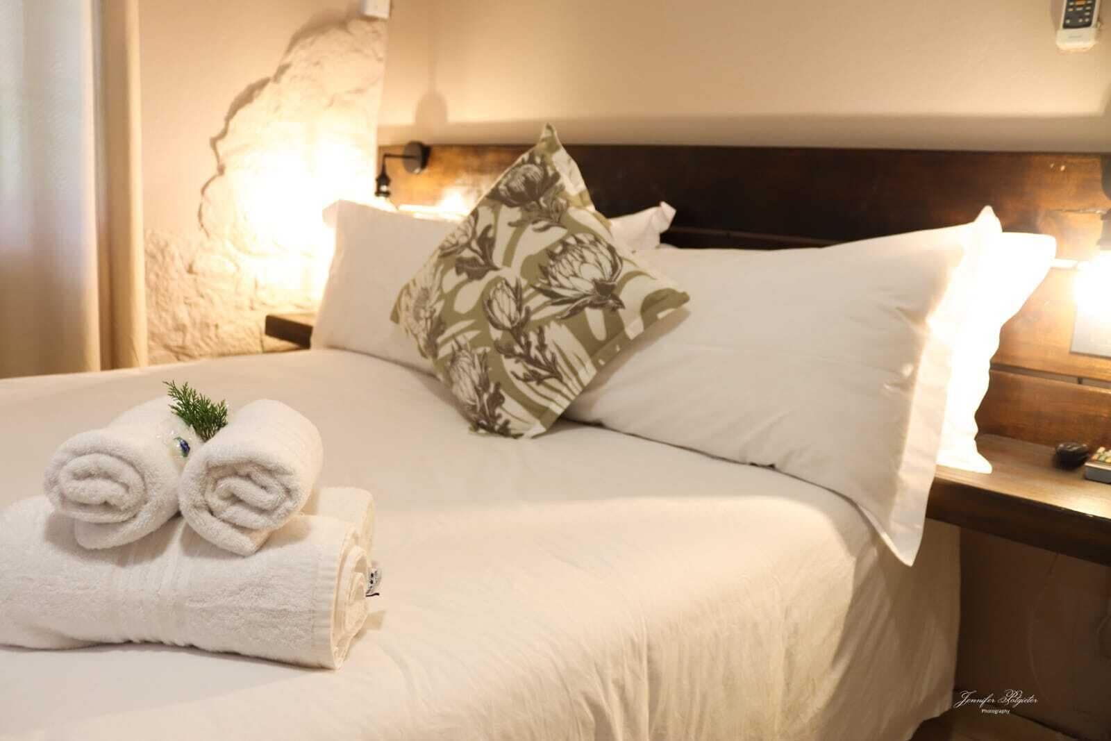 Una cama bien hecha con sábanas blancas, un cojín floral y toallas enrolladas al lado.