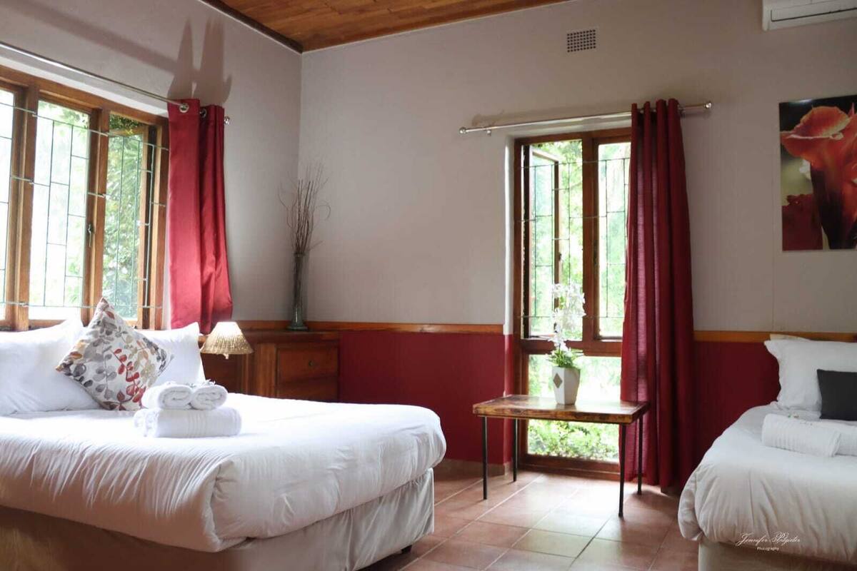A hotel room with two beds, a small table, a window with red curtains, and a potted plant.