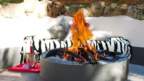 A fire pit with burning logs, surrounded by a stone wall and a zebra-patterned cushion. - The Ultimate Lodge (Louis Trichardt)