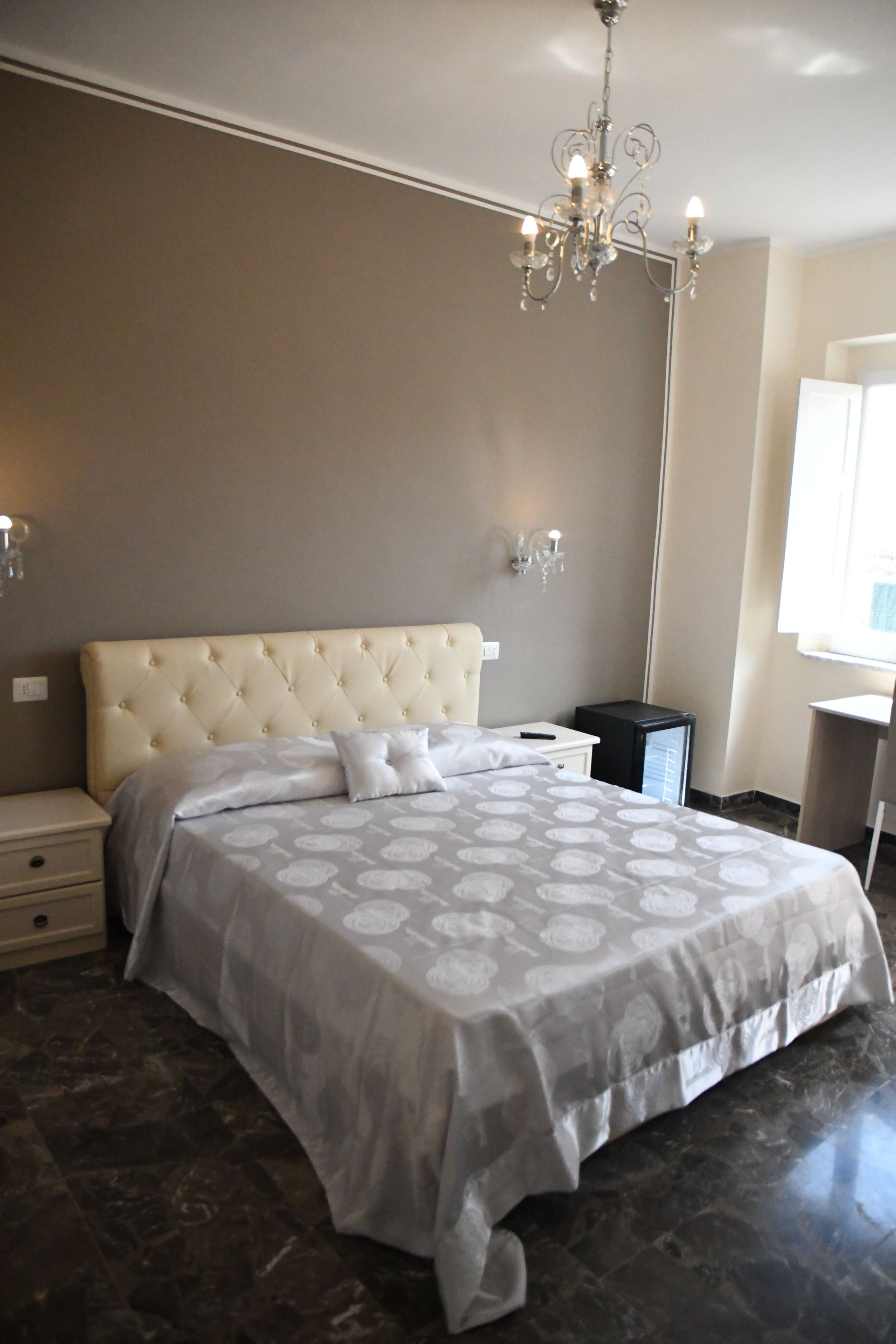 A bedroom with a tufted headboard, a large bed with a patterned bedspread, a bedside table, a nightstand, a desk, a chair, and a chandelier.
