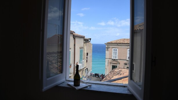 A window with a bottle and glass on the sill, overlooking a coastal street with buildings and a clear sky. - B&B THE TOWER Tropea (Tropea)