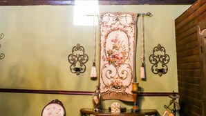 Una habitación con techo alto, vigas de madera a la vista, una araña, un tapiz decorativo, un sillón acolchado, una mesita con un florero y un perchero.