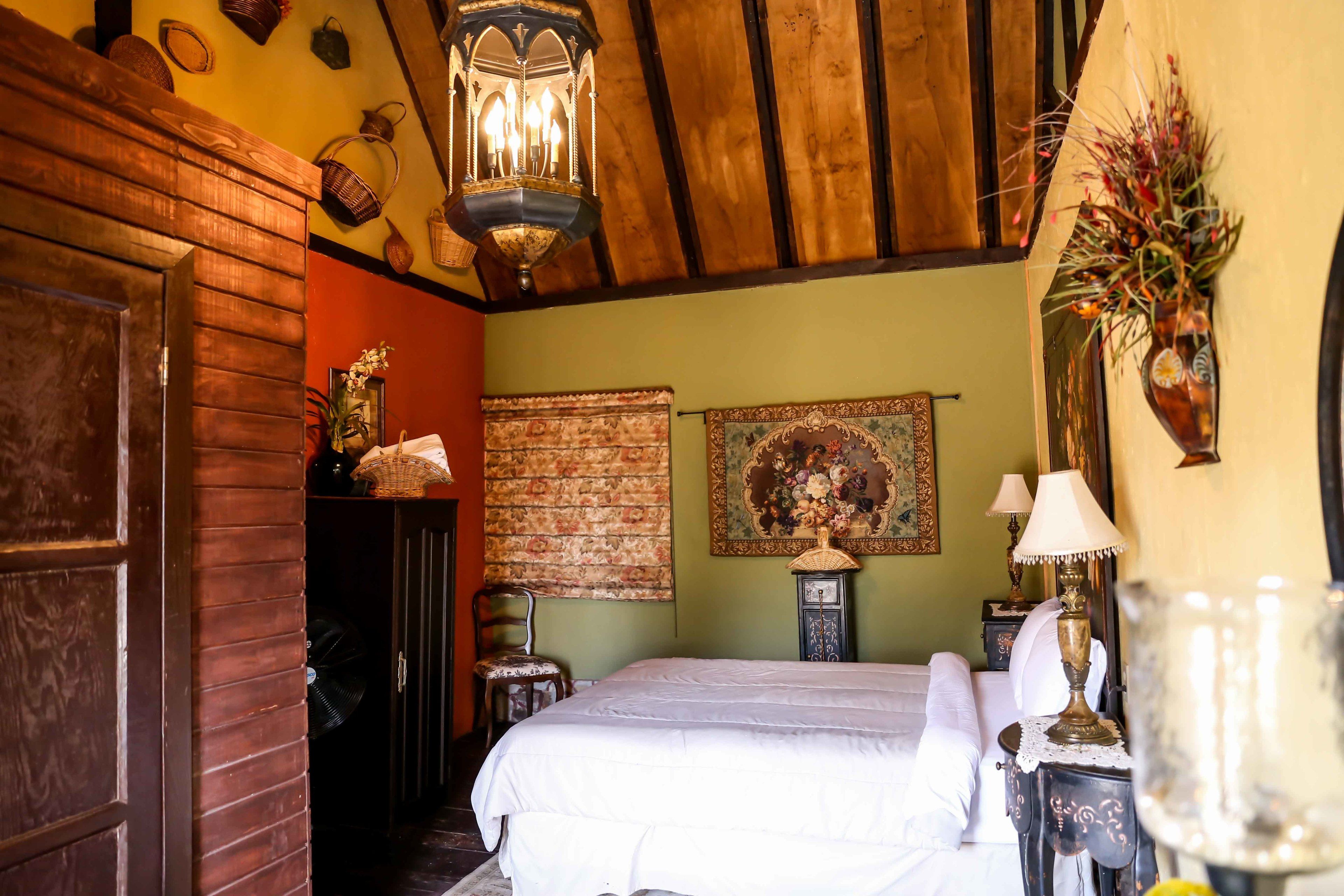 A cozy room with a wooden ceiling, a bed with white linens, a wooden door, and a wall with a tapestry and a lamp.