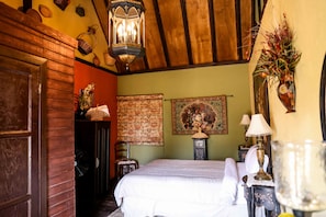 A cozy room with a wooden ceiling, a bed with white linens, a wooden door, and a wall with a tapestry and a lamp. - Fernabella's Lodge (Valle de Guadalupe)