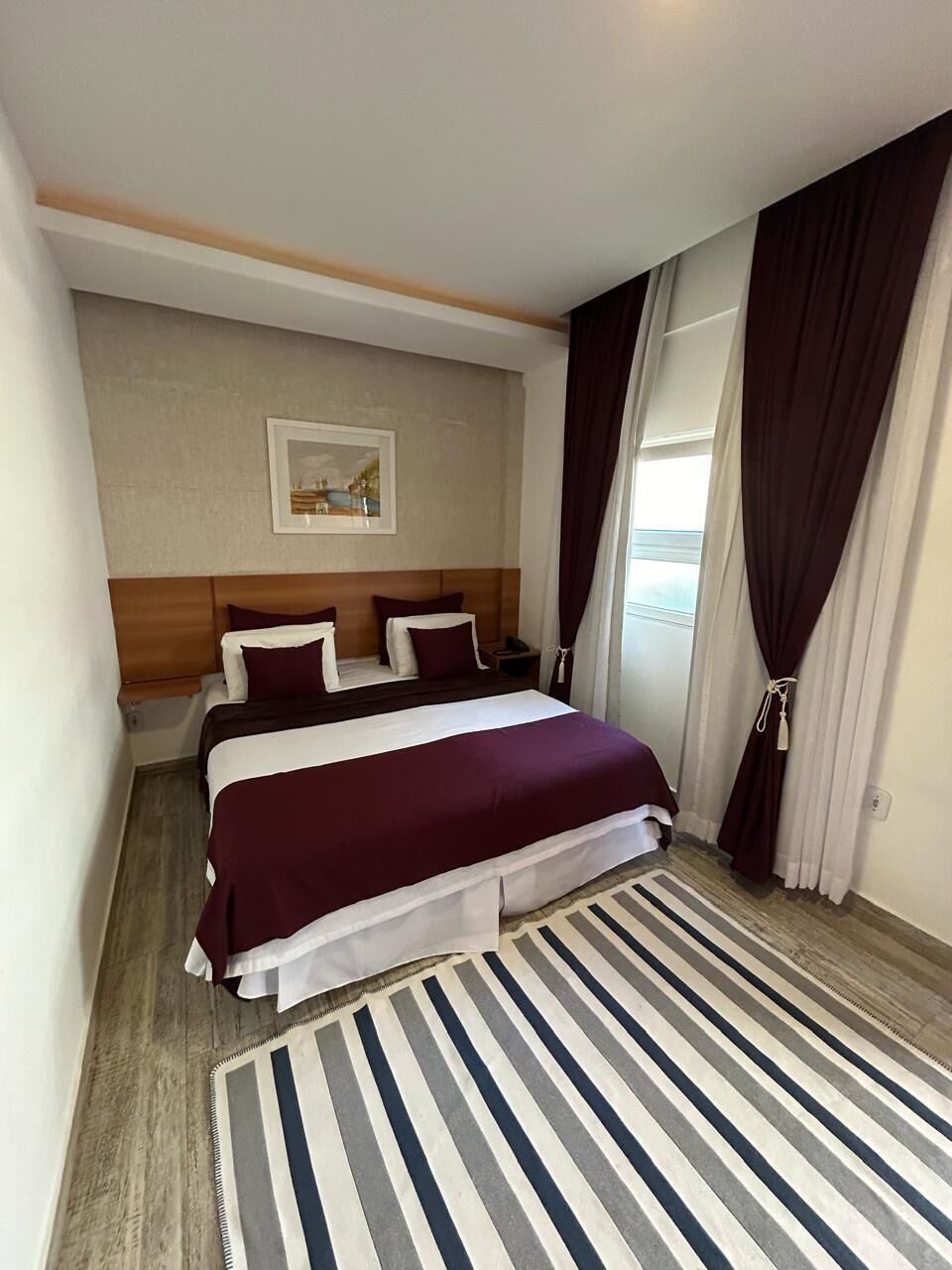 A hotel room with a bed, a nightstand, a painting on the wall, and a view of the outdoors.