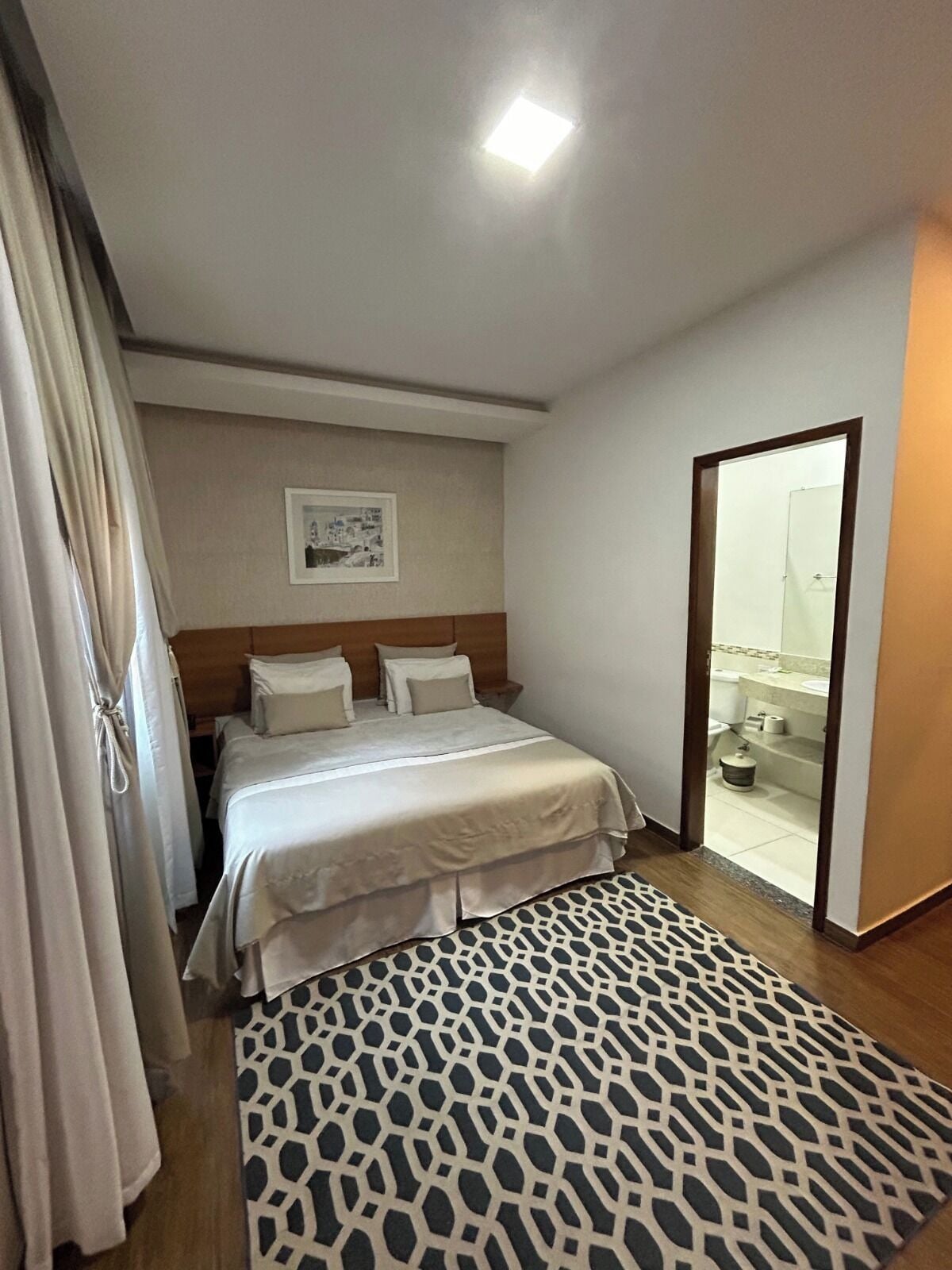 A hotel room with a bed, a patterned rug, a framed picture on the wall, and a bathroom visible through an open door.