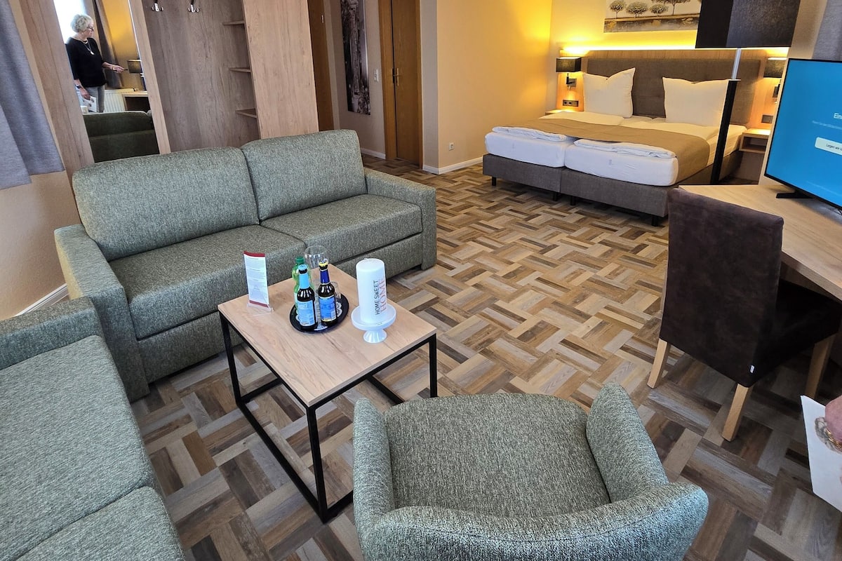 A modern hotel room with a sofa, armchair, and a bed. There is a small table with drinks and a candle.