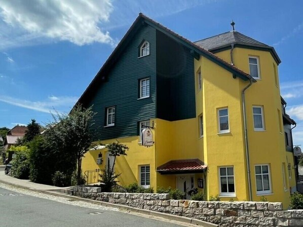 Un edificio de dos plantas con fachada verde y amarilla, un muro de piedra en primer plano y un cielo azul despejado al fondo.