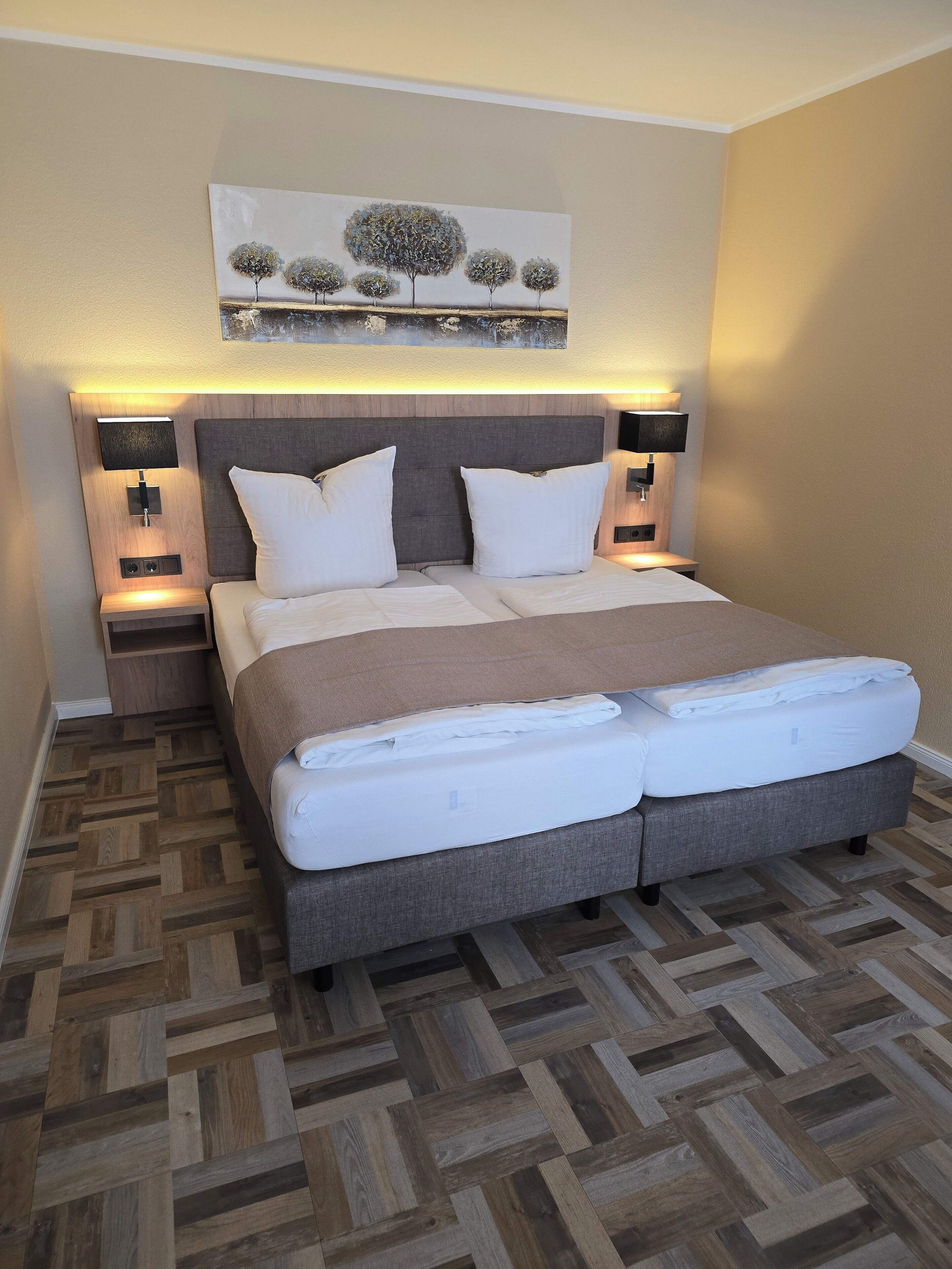 Una cama bien hecha con cabecera gris, dos mesitas de noche con lámparas, y un cuadro sobre la cama.