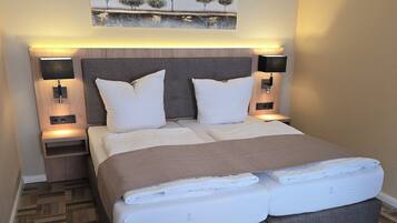 Una cama bien hecha con cabecera gris, dos mesitas de noche con lámparas, y un cuadro sobre la cama.