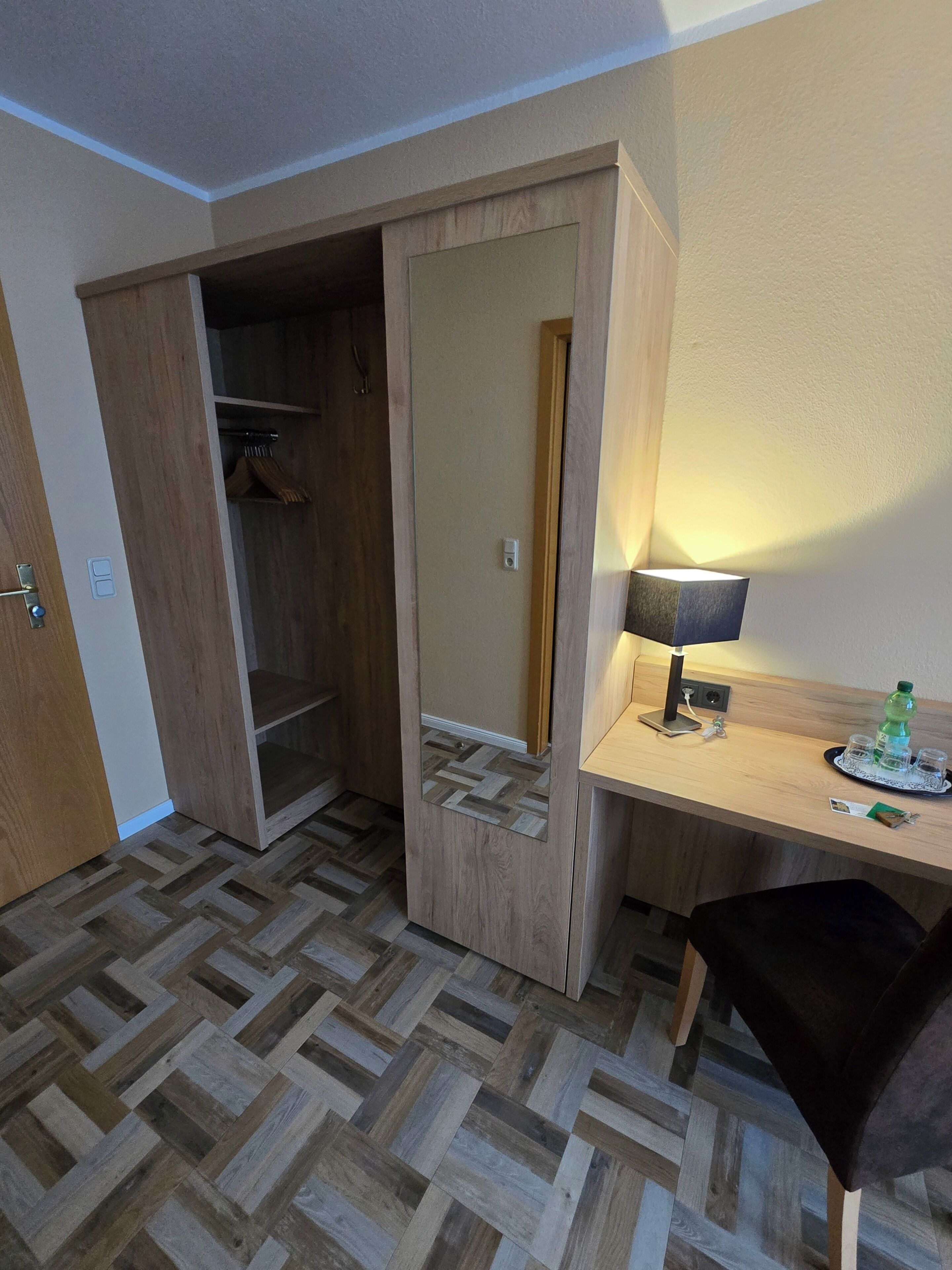 Ein Hotelzimmer mit einem Holzschrank, einem Schreibtisch mit Lampe, einem Stuhl und einem Fiskgrätparkettboden.